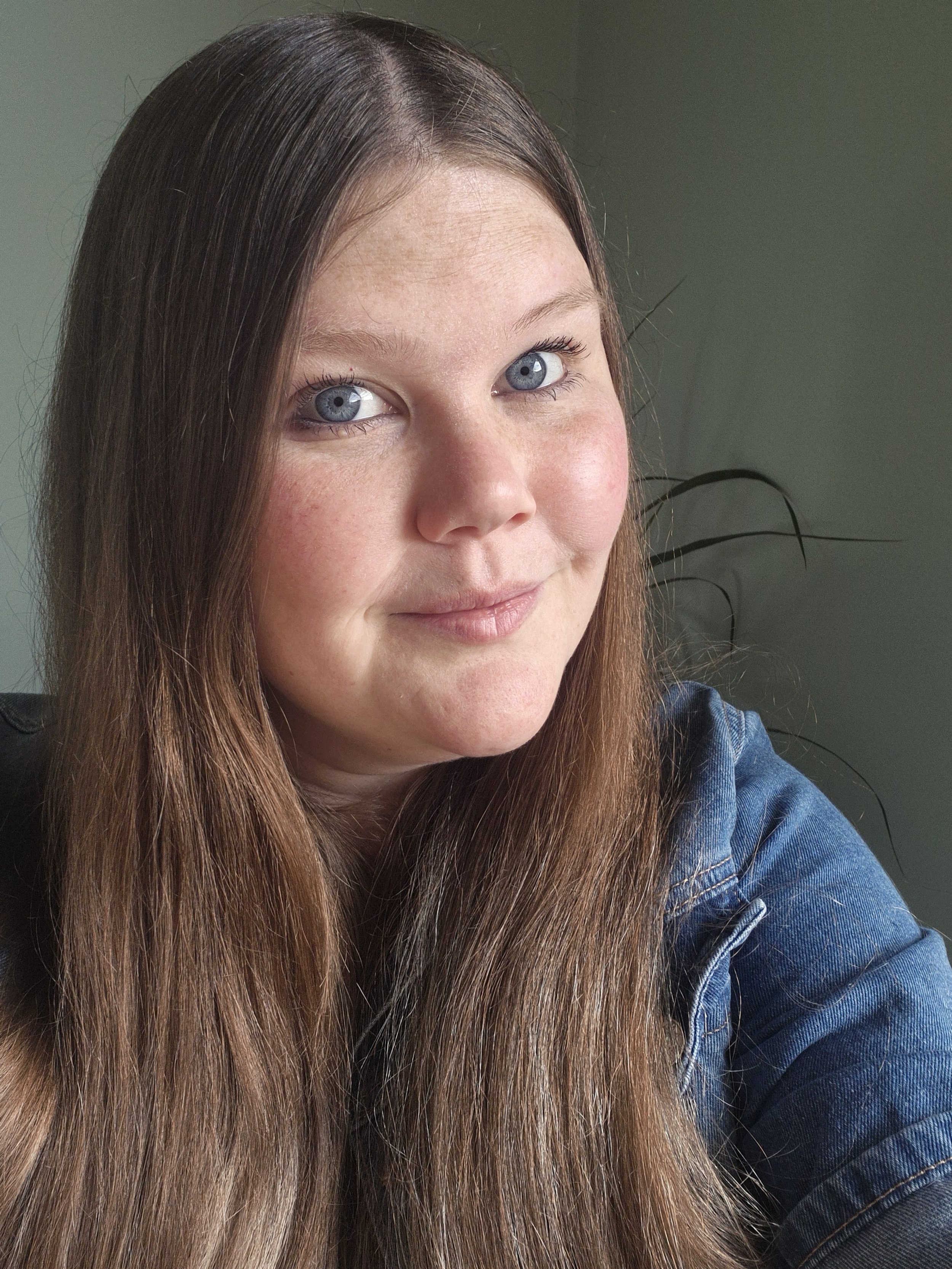 Smiling person with long brown hair and blue eyes wearing a denim jacket.