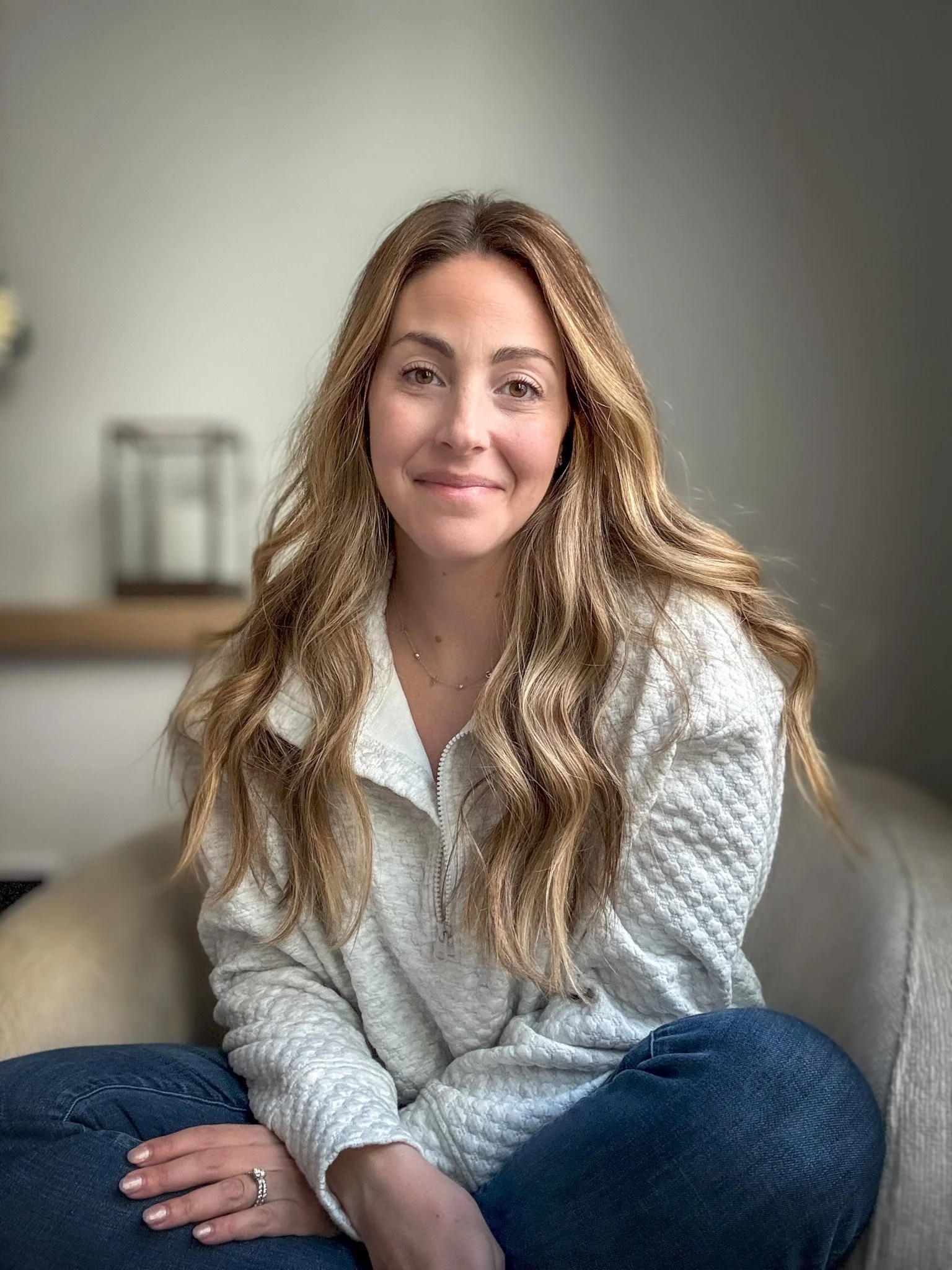 A woman with long wavy hair sitting on a beige sofa, smiling at the camera, wearing a white zip-up sweater and blue jeans.