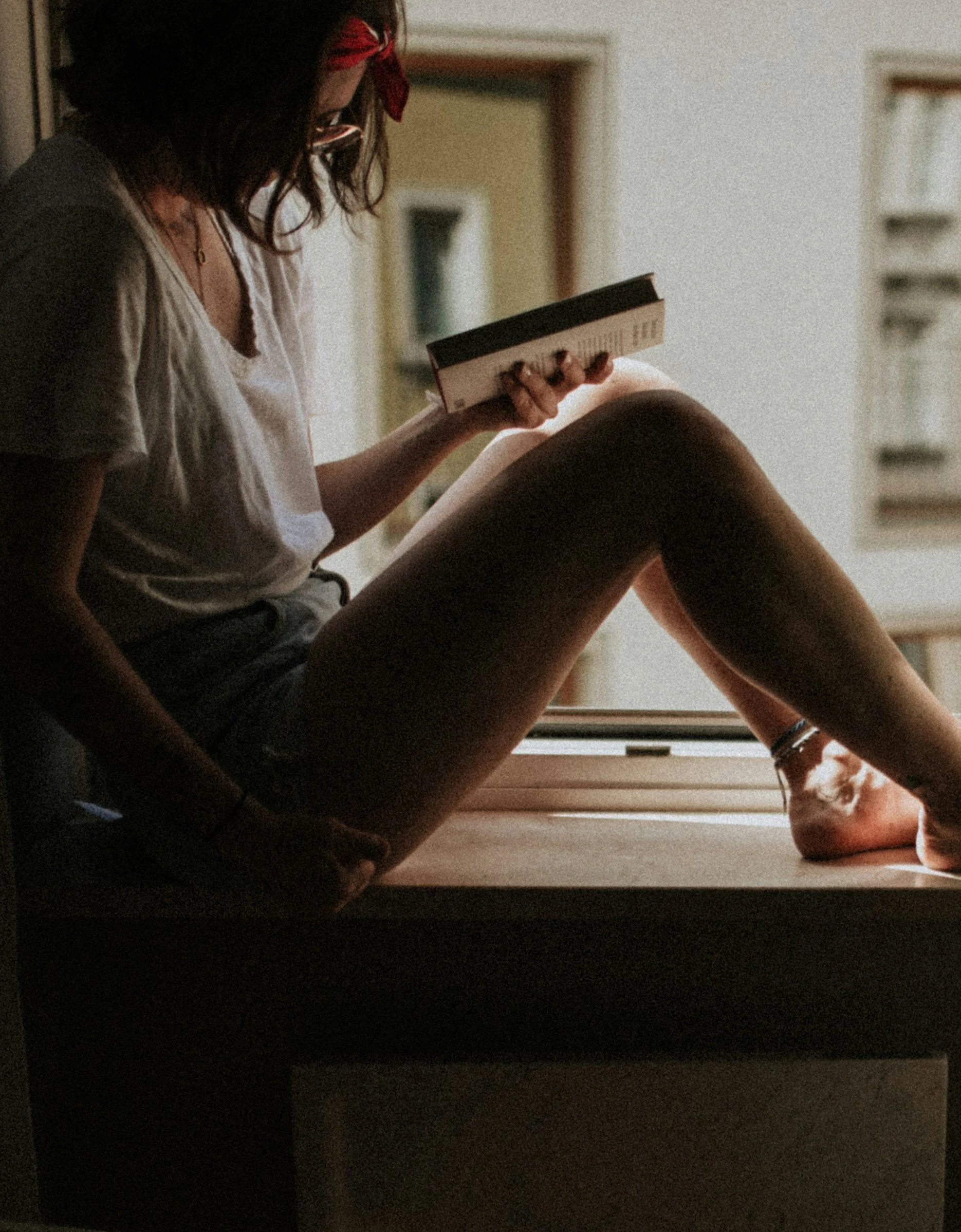 A person sitting on a windowsill reading a book, with their legs extended and bent at the knees. The person appears to be in a cozy indoor space with bookshelves in the background.