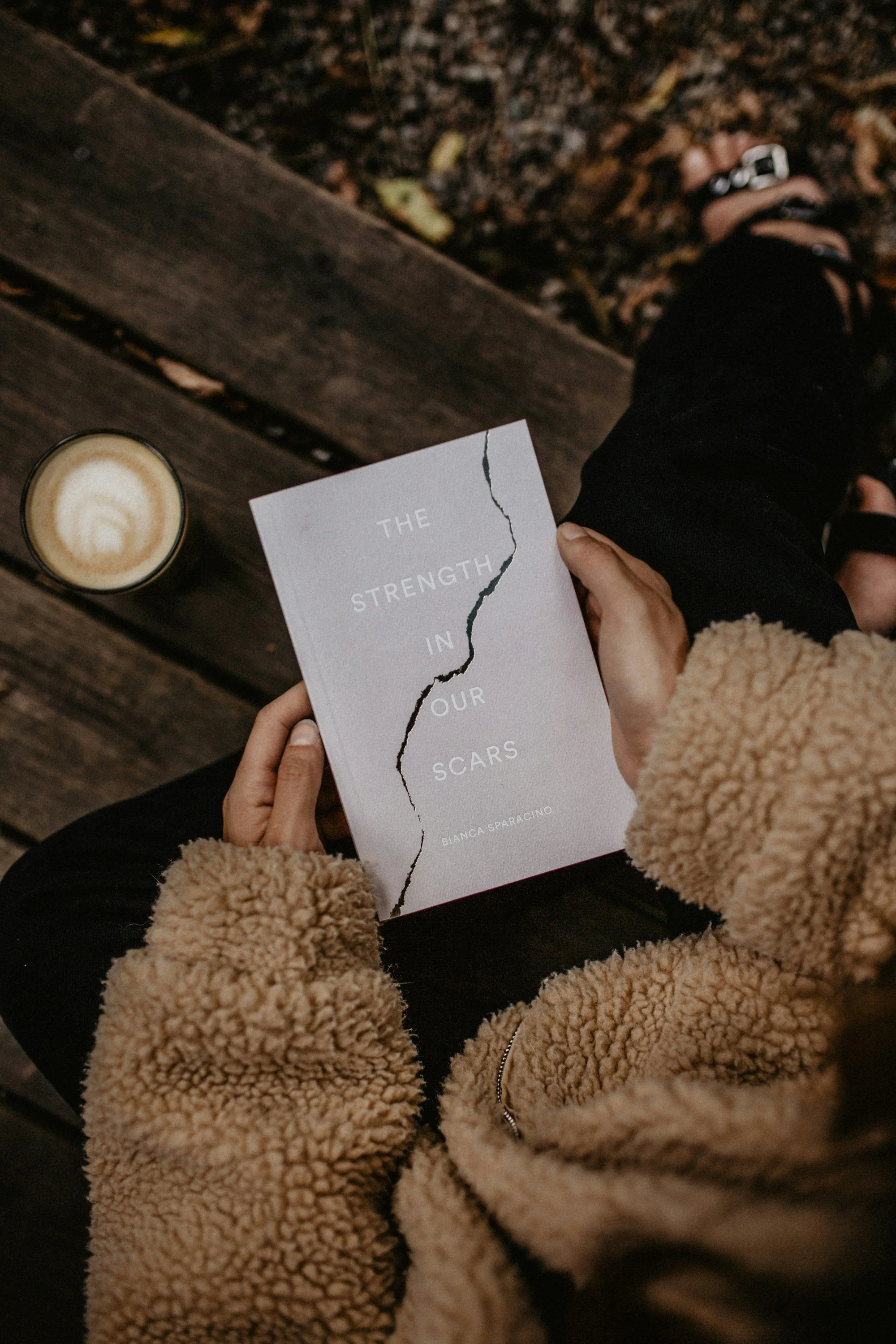 Person sitting outdoors on a wooden bench, wearing a tan fuzzy jacket and black pants, holding a book titled 'The Strength in Our Scars' by Bianca Sparacino, with a latte on the bench beside them.
