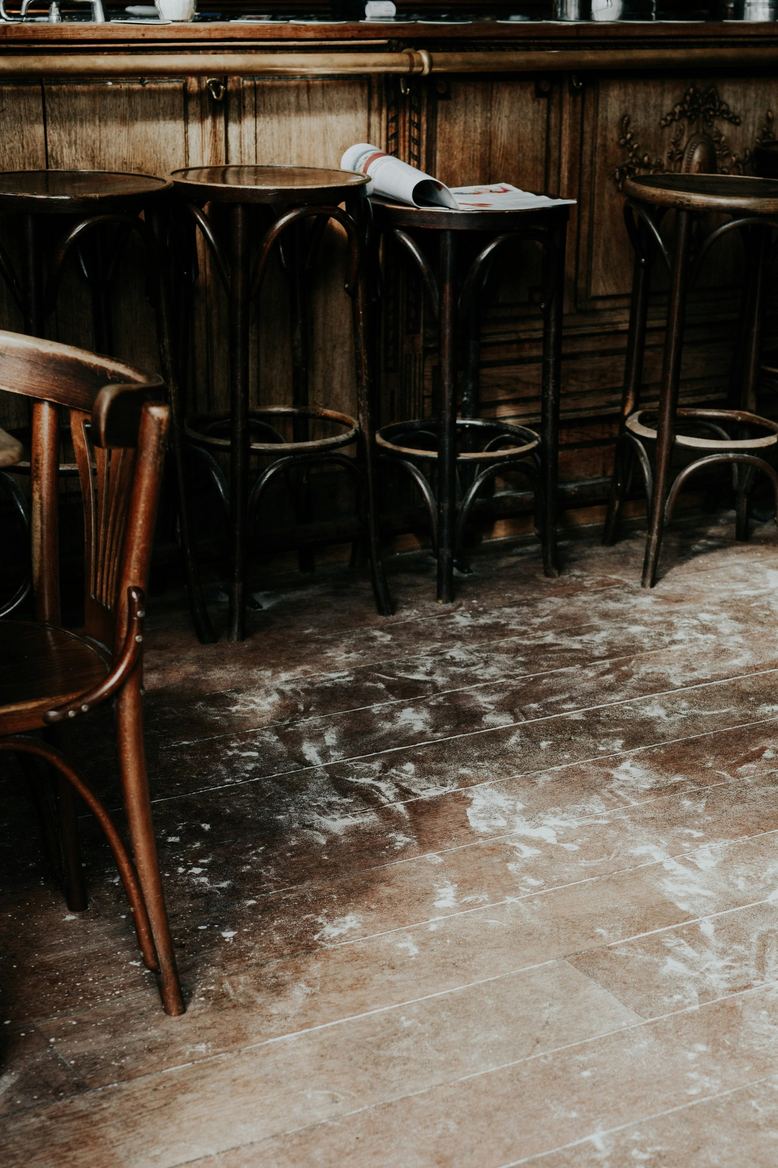 Dirty wooden floor inside a rustic cafe or bar with dark wooden tables and chairs, some of which are overturned or placed upside down on the tables.
