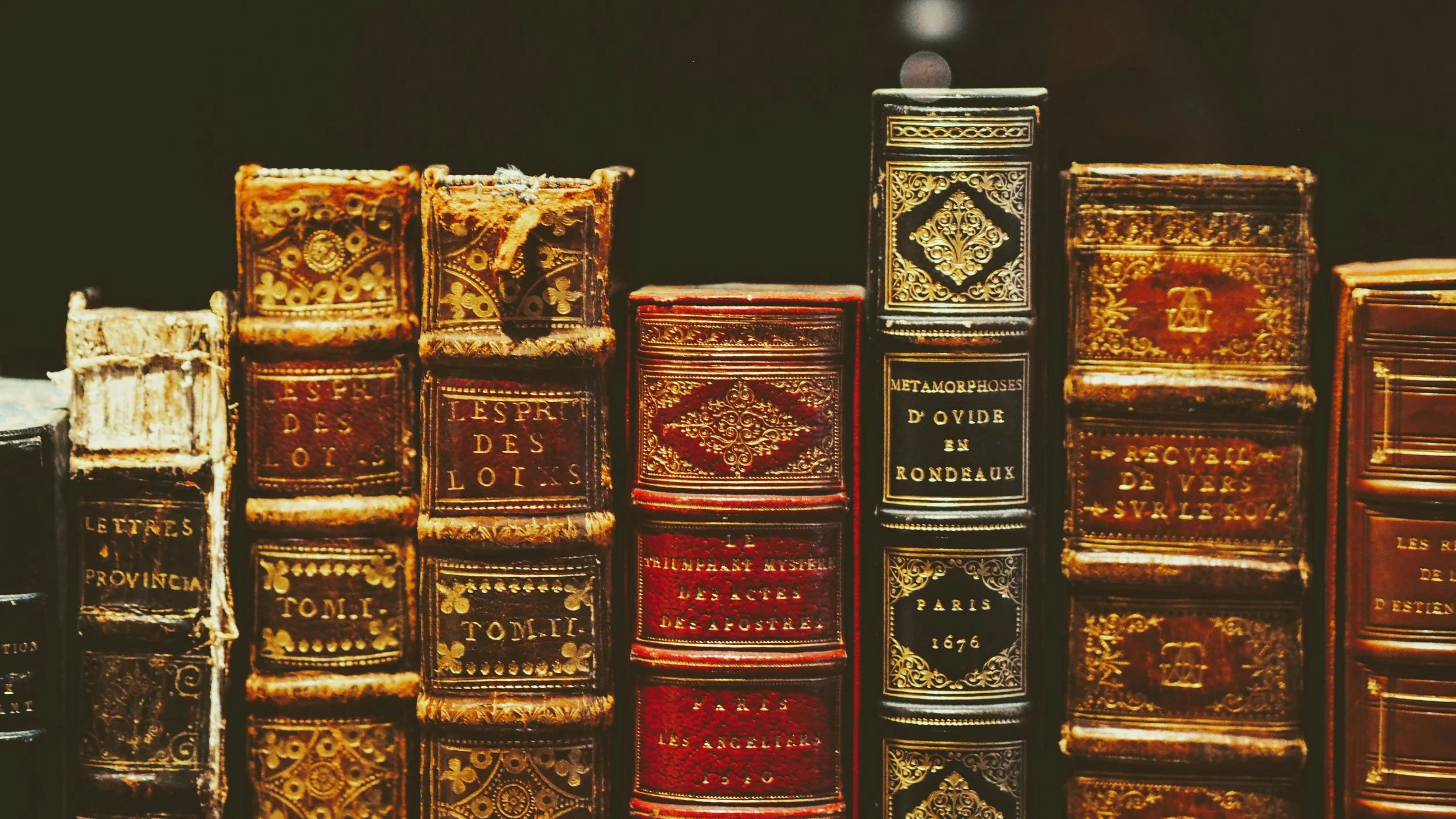 Close-up of antique, ornate leather-bound books on a shelf, with gilded titles and intricate gold embossing.