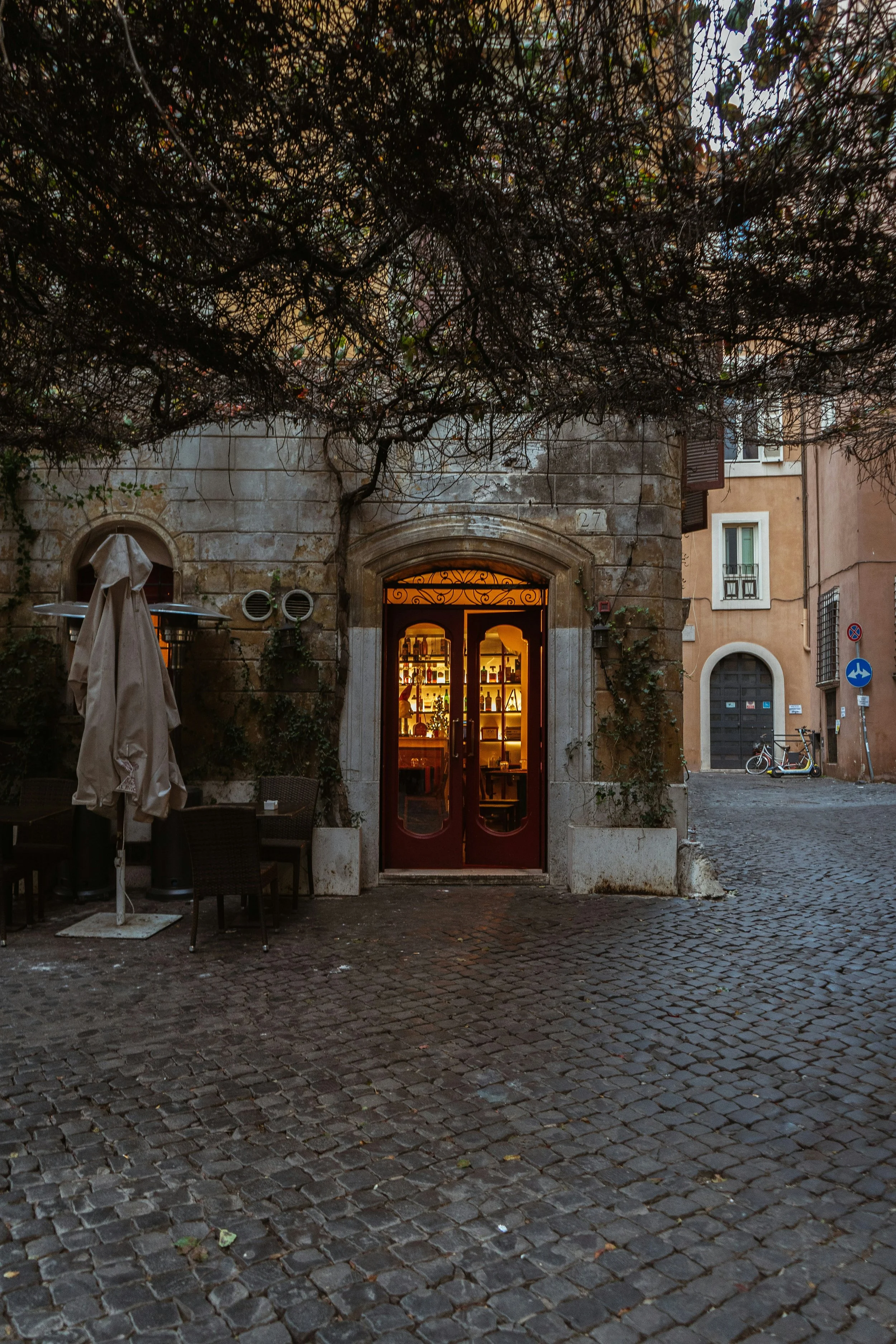 An outdoor scene of a cobblestone street at dusk with a small restaurant or bar entrance, illuminated from within, with chairs and a folded umbrella outside, and a bicycle leaning against a wall in the background.