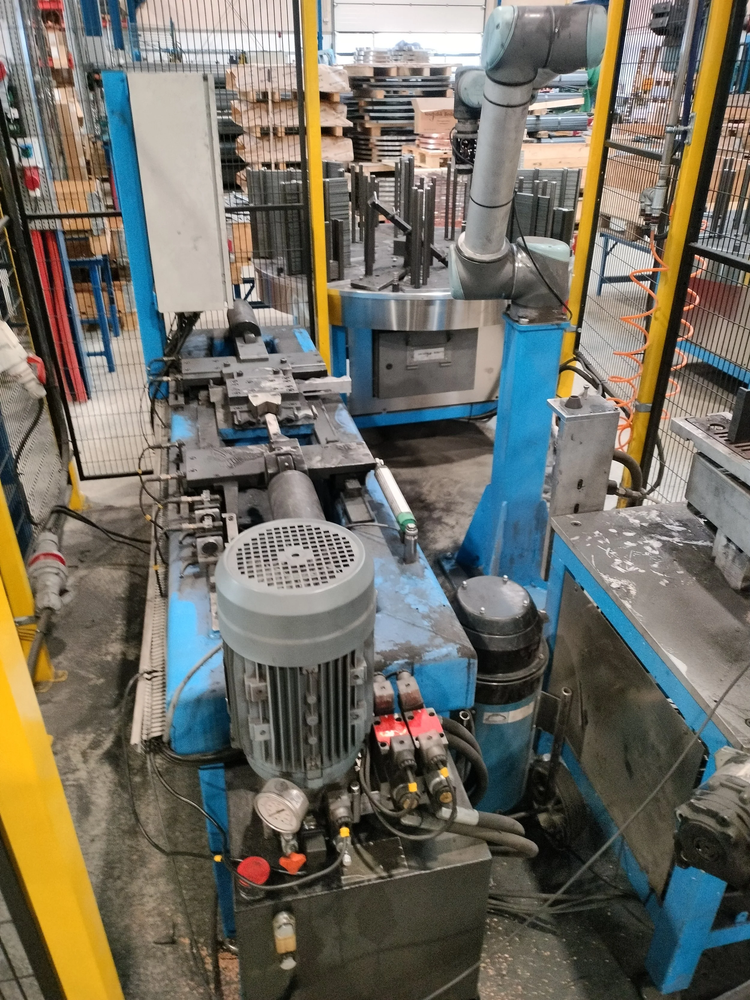 Industrielt maskineri og udstyr i en fabrikshal, herunder en maskine med en stor elektrisk motor og forskellige tilslutninger, samt andre værktøjer og materialer i baggrunden.
