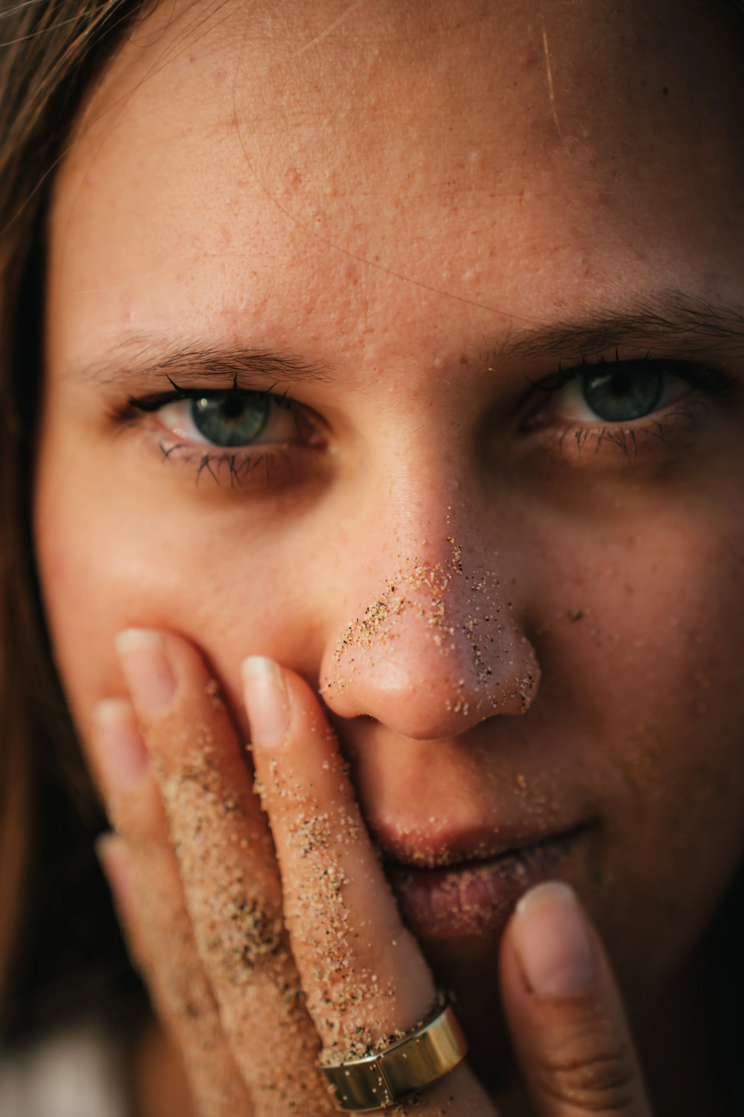 Nahaufnahme eines Gesichts mit Sand an Nase, Lippen und Hand, blaue Augen, braune Haare und goldener Ring am Finger.