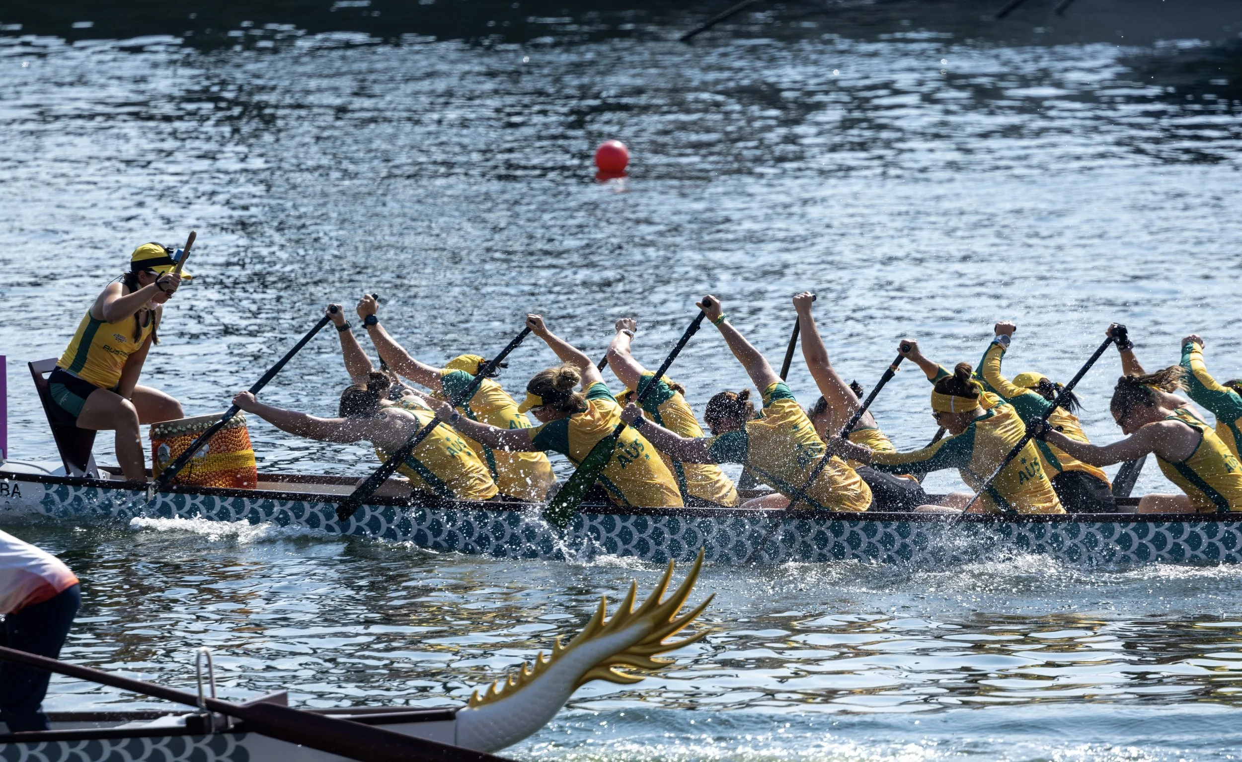 The national Australian dragon boat team competing in a dragon boat race on the water, wearing yellow and green uniforms, paddling in unison.