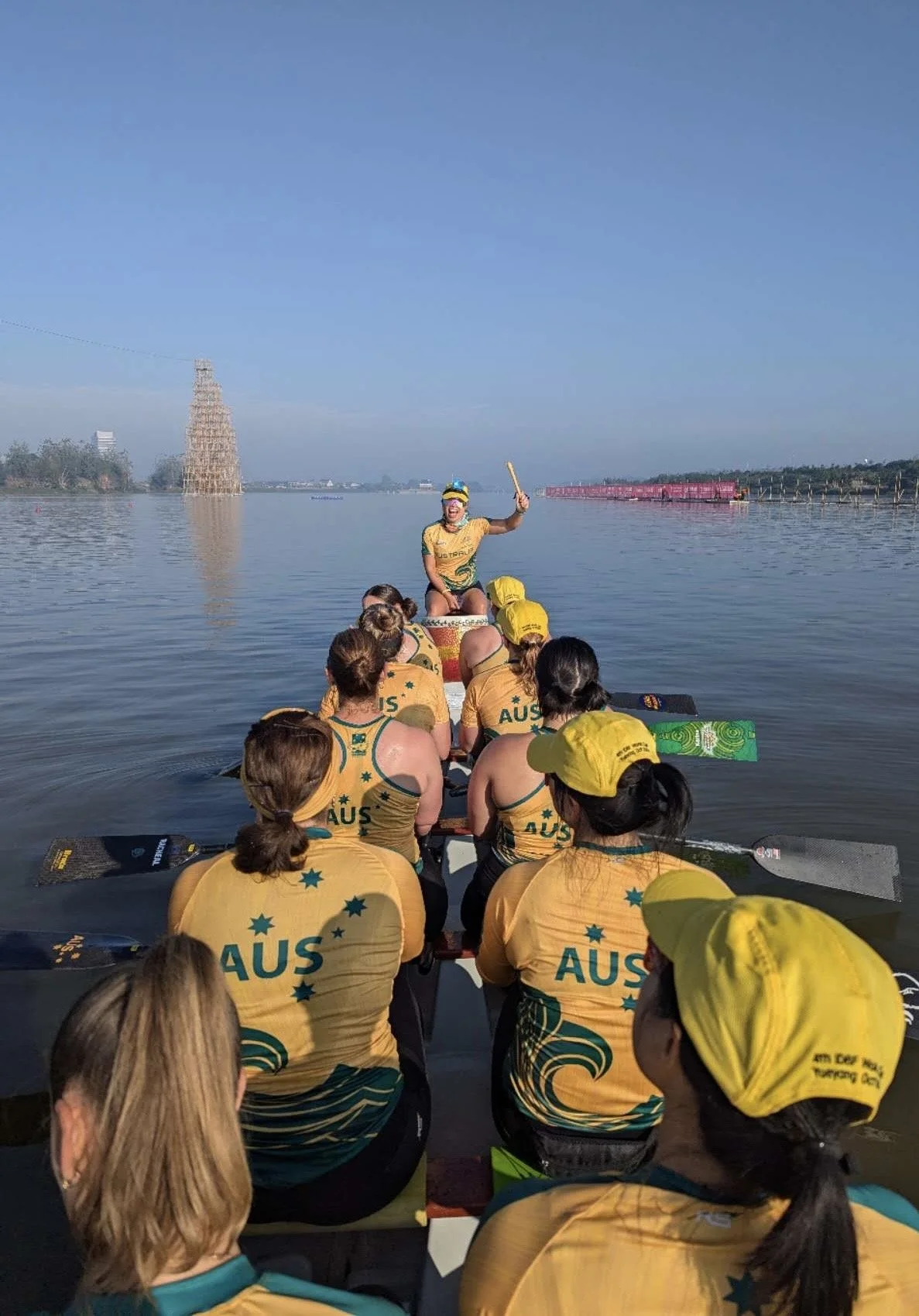 A group of women in yellow Australian team sports uniforms with 'AUS' on the back, sitting in a dragon boat on calm water, with a woman in front holding a drum and raising a stick, leading the team.