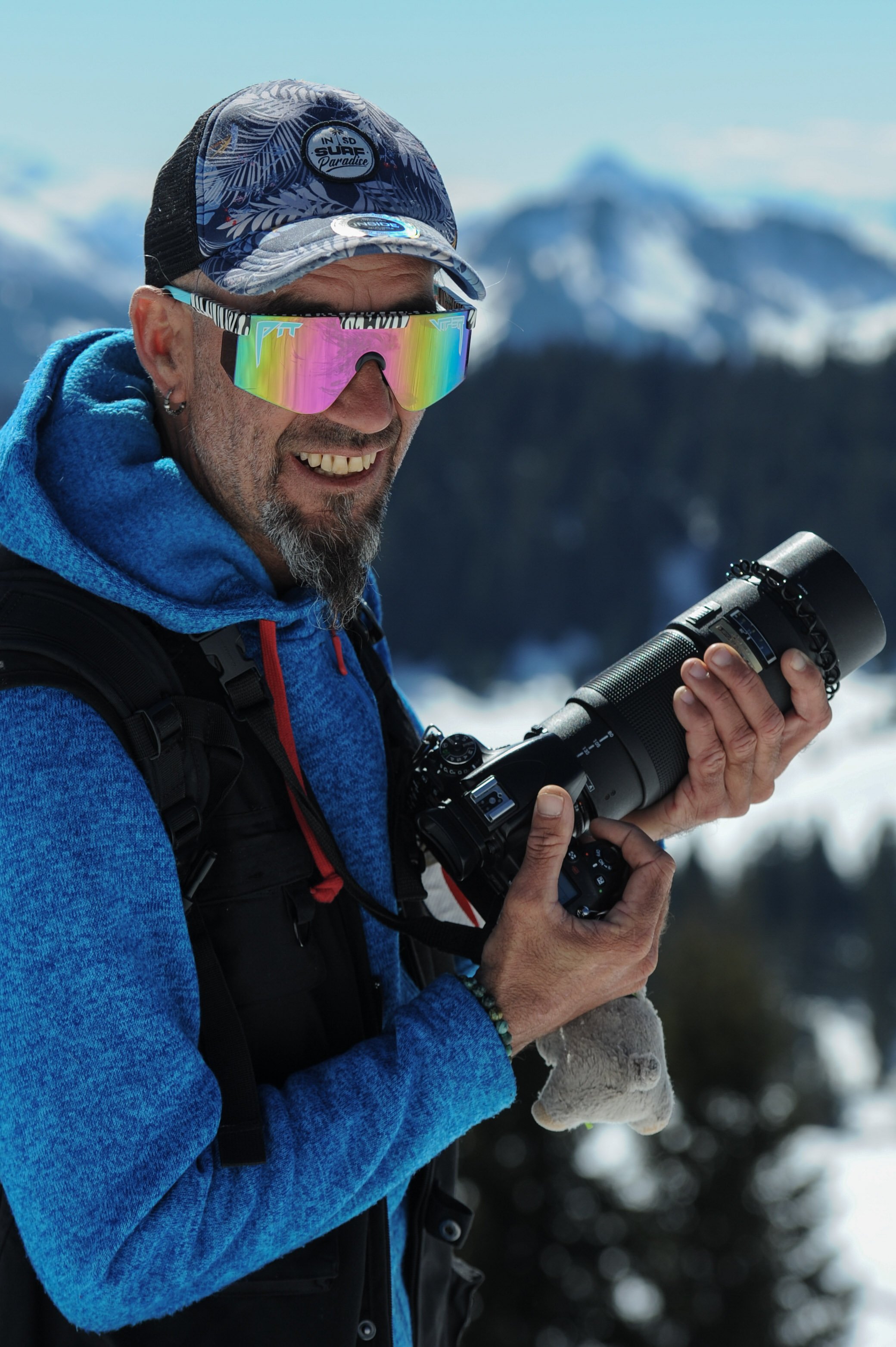 Homme portant des lunettes de soleil réfléchissantes et un hoodie bleu, tenant un appareil photo avec un objectif long, devant un paysage de montagne enneigé.