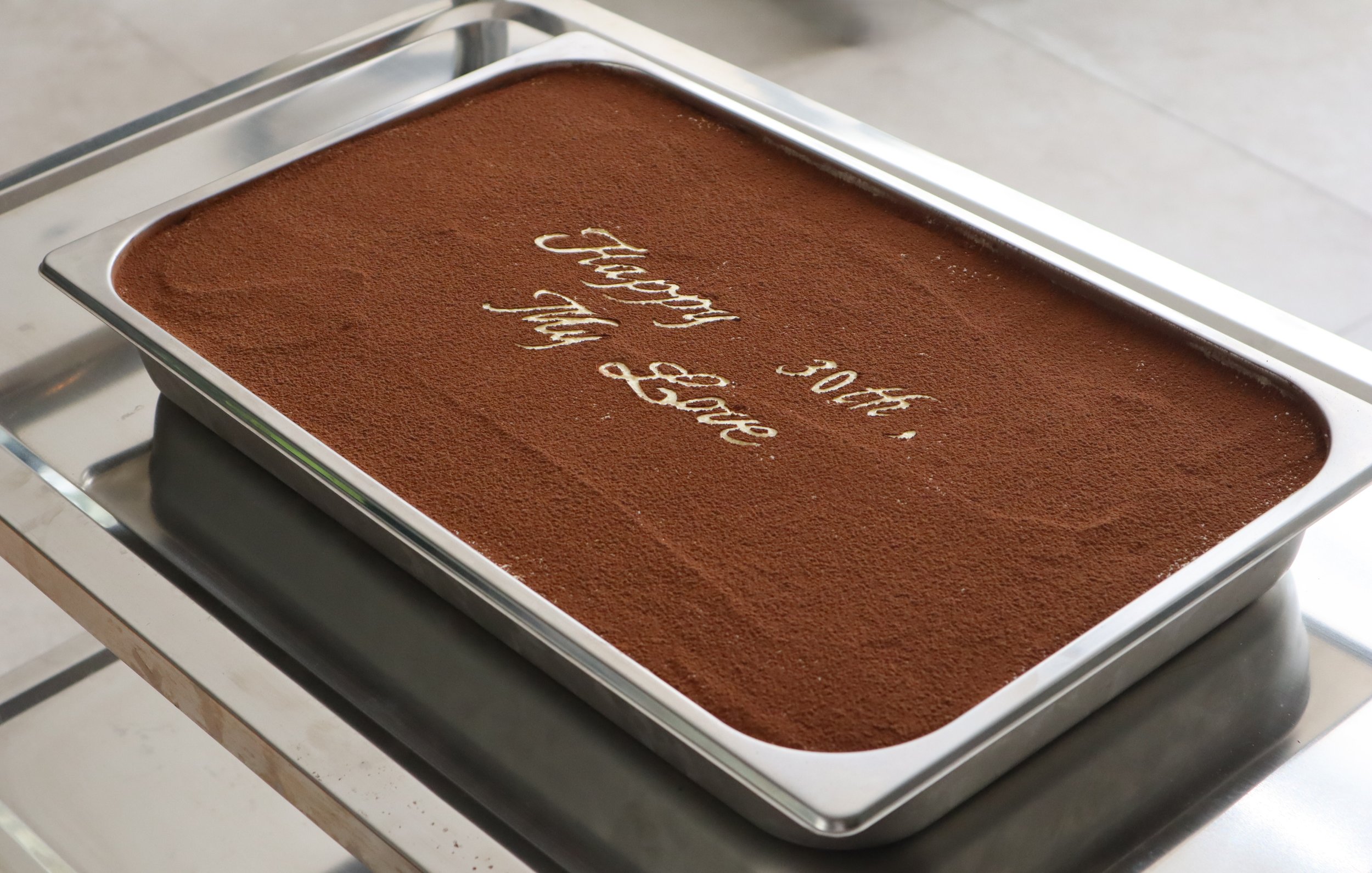 A tray of tiramisu with cocoa powder and the words "Happy 30th, My Love" written on top.
