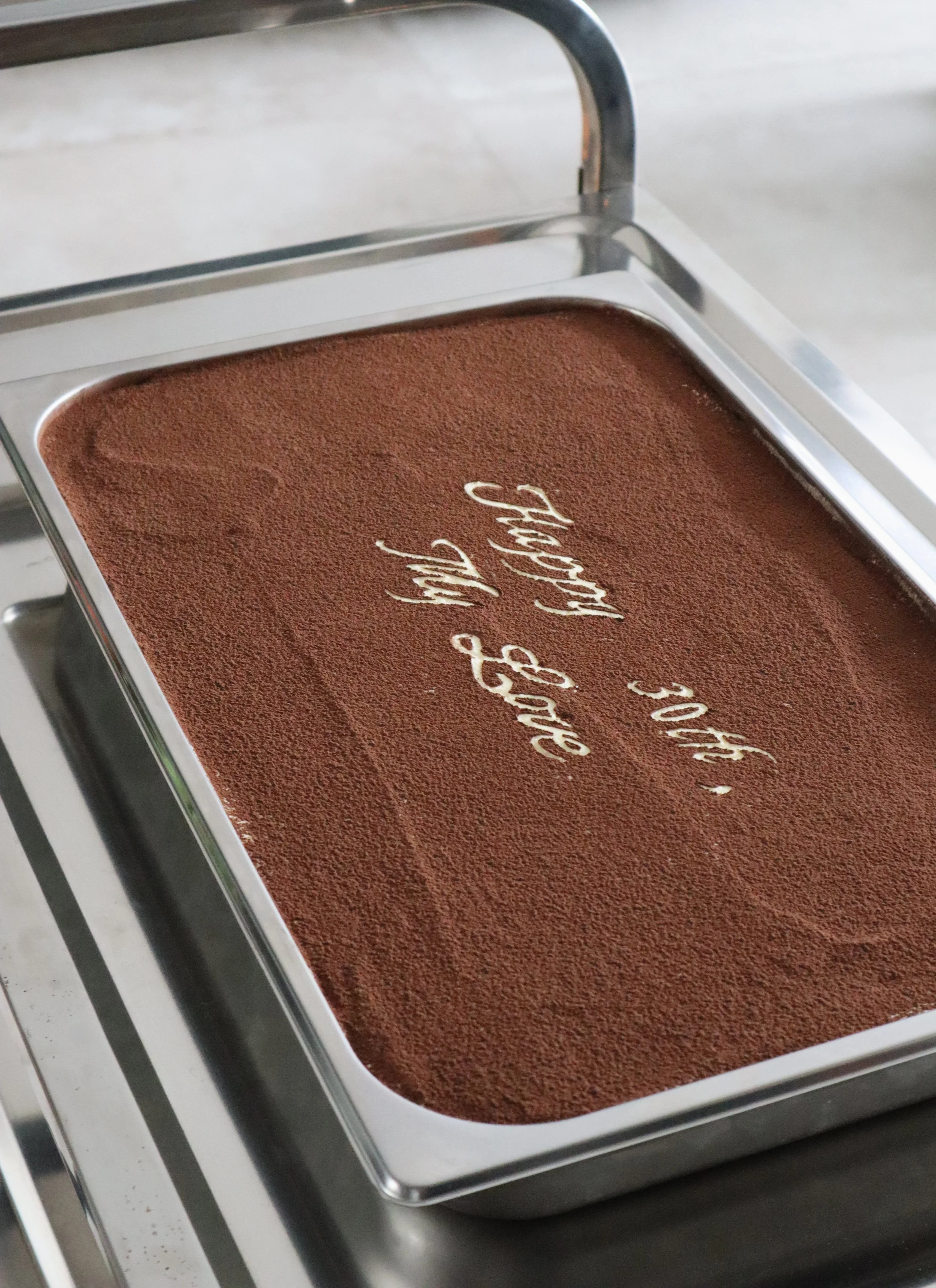 A rectangular stainless steel tray filled with tiramisu layered dessert, topped with cocoa powder and decorated with white chocolate writing that says "Happy 30th, My Love".