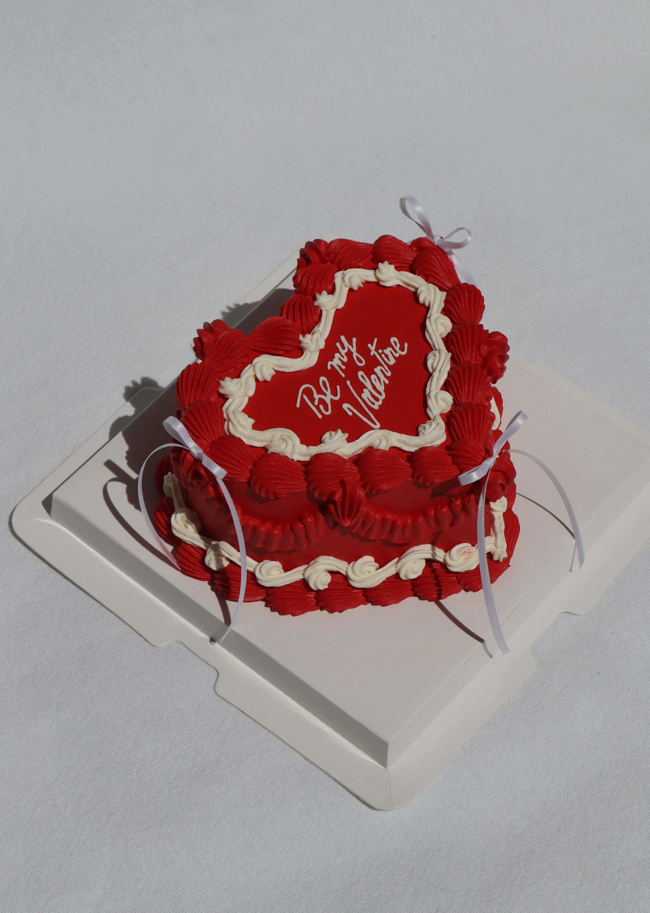 Heart-shaped cake with red and white icing, decorated with ribbons and piping, with the message 'Be My Valentine' written on top.