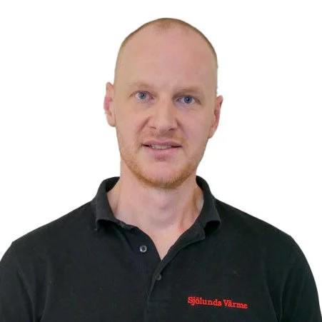 En man med nästan helt rakat huvud, blå ögon och lätt skägg, som bär en svart polo med röd text 'Sjölundas Värme' på bröstet, mot en vit bakgrund.