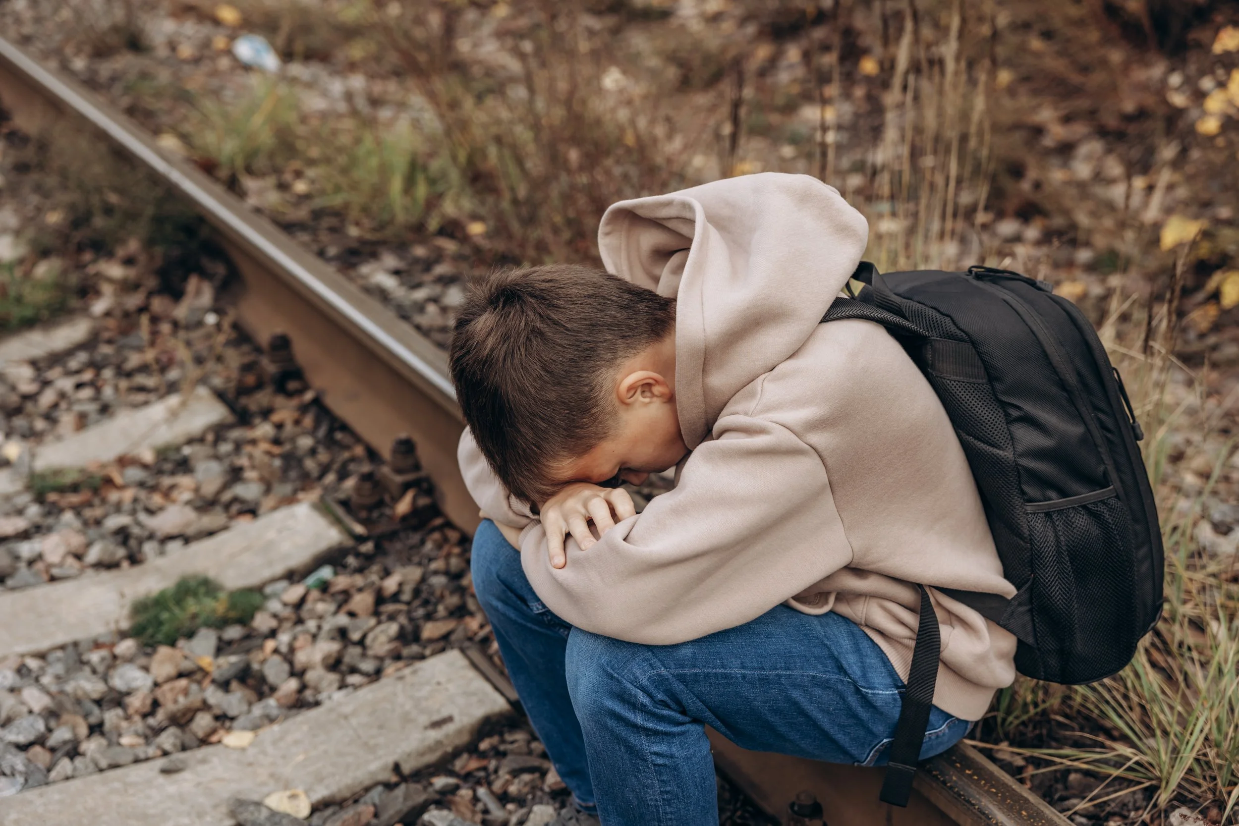 A person sitting by railroad tracks with their head resting on their arms, wearing a beige hoodie and a black backpack.