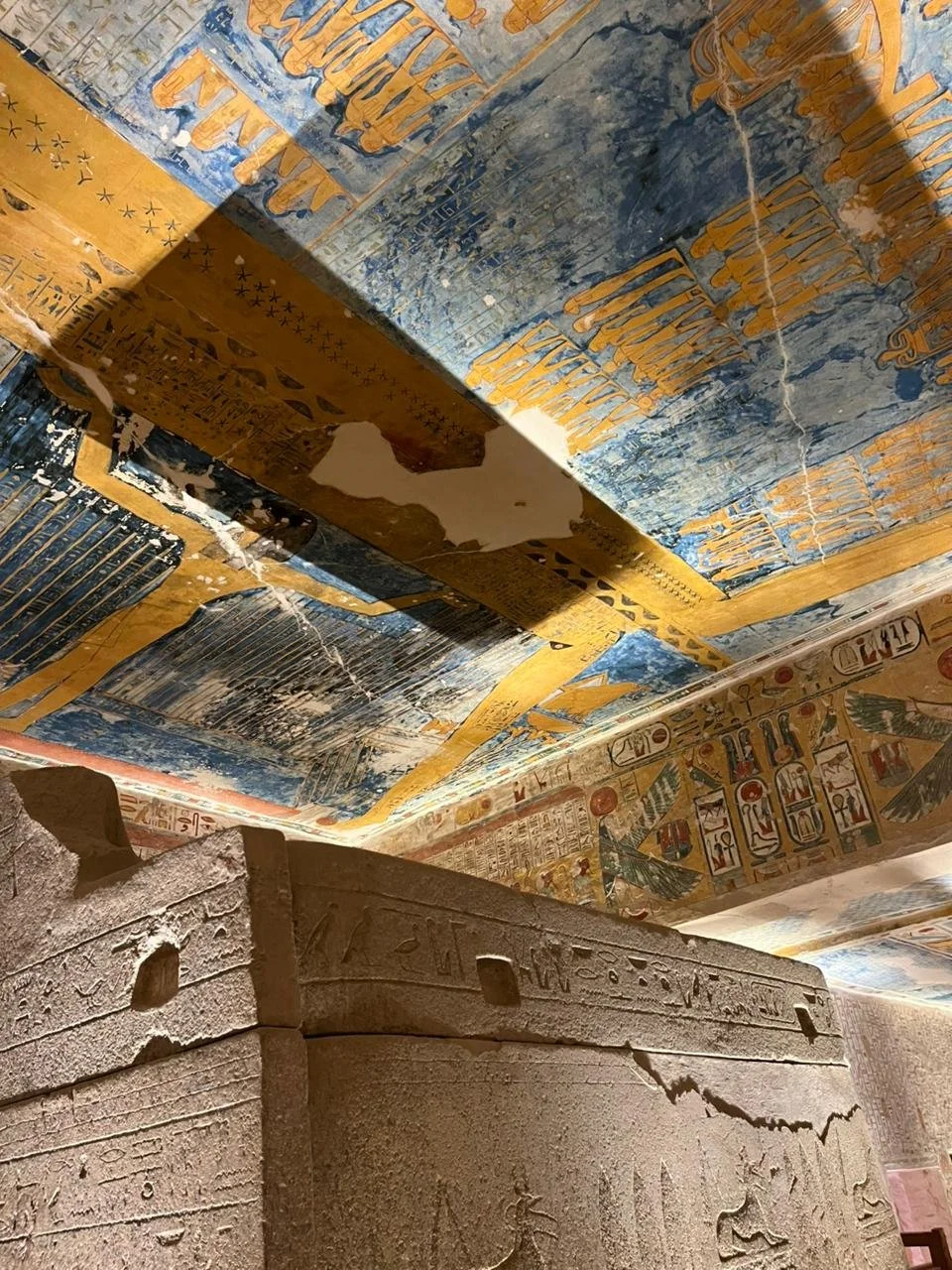 Ancient Egyptian hieroglyphic carvings on stone walls and a painted ceiling with hieroglyphs and symbols in blue, gold, and red, indicating an Egyptian tomb or temple interior.