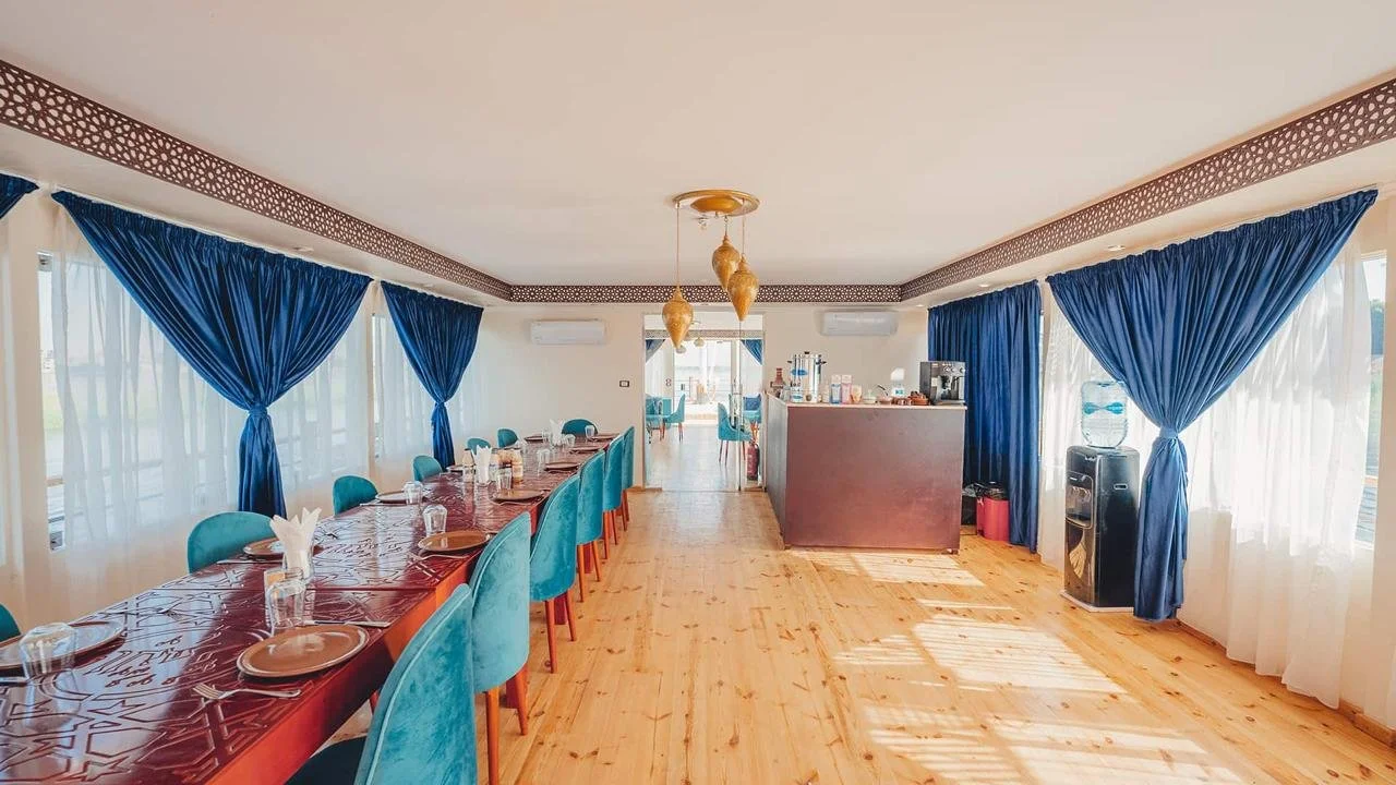 A dining room with a long table set with plates, glasses, and napkins, surrounded by blue chairs. The room has large windows with dark blue curtains, a water dispenser, and a counter with coffee machines, all illuminated by natural light.