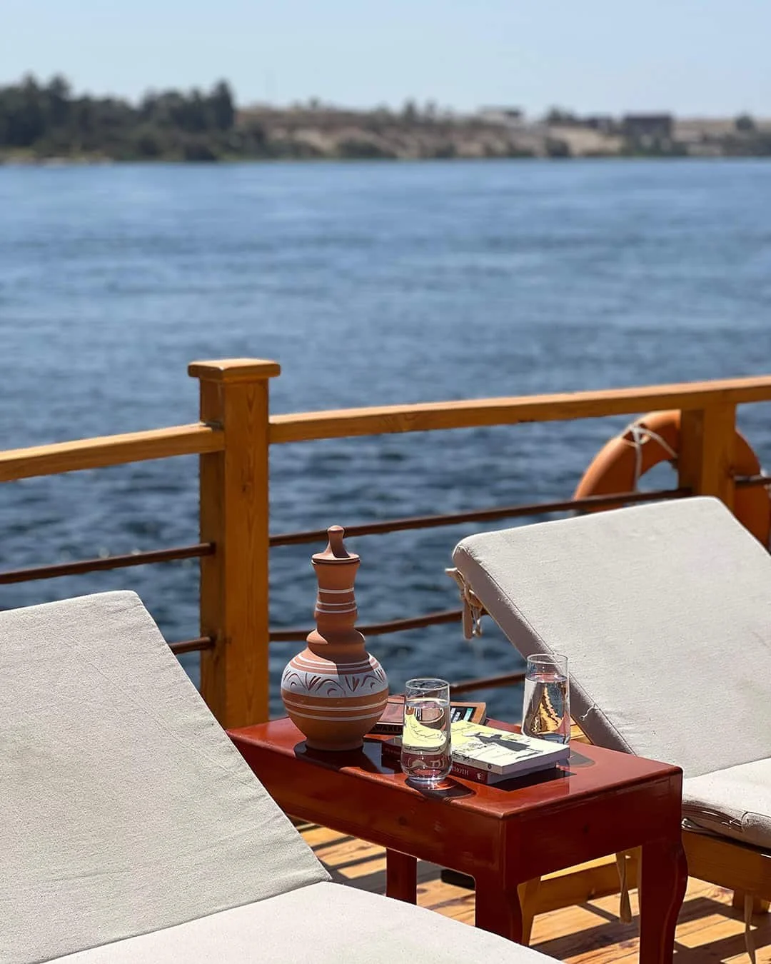 A view from a boat deck with two white lounge chairs, a small wooden table, a decorative ceramic vessel, and two glasses of water, overlooking a body of water with land in the distant background.