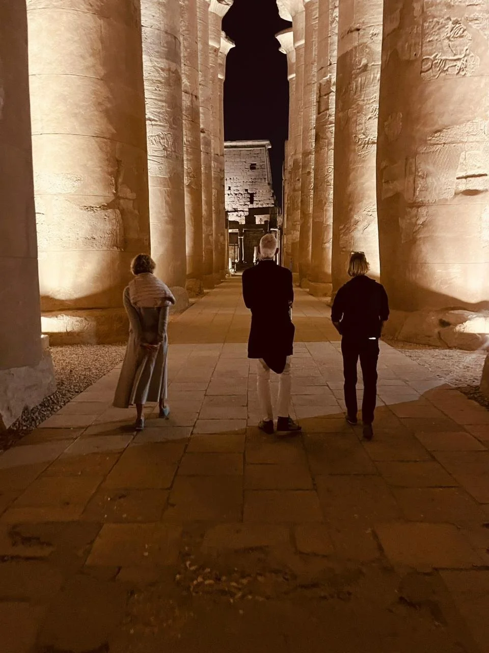 Three people standing among ancient, illuminated columns at night in an archaeological site.