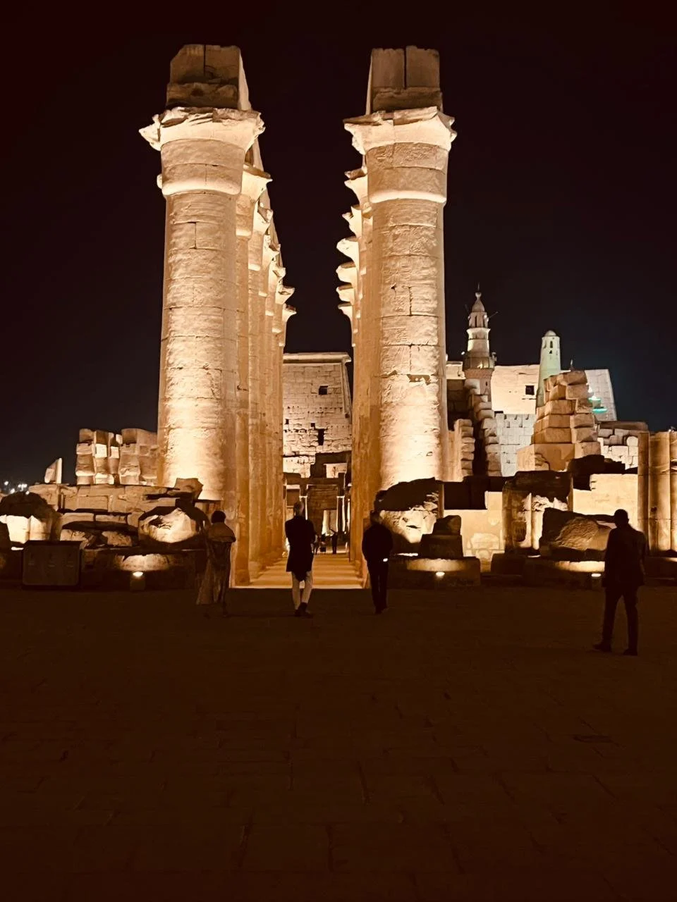 Ancient Egyptian temple ruins lit at night, with large stone columns and a few people walking through.