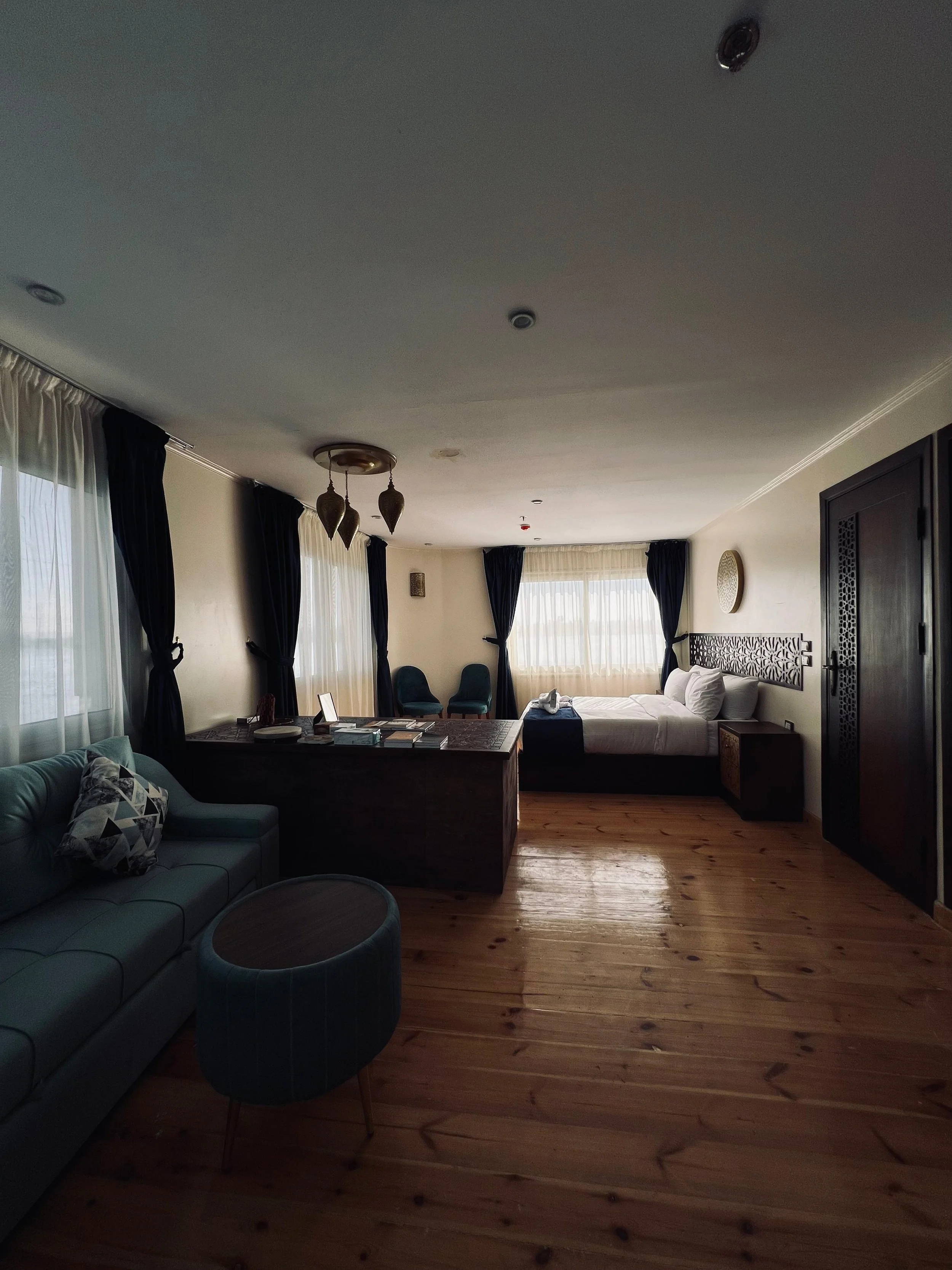 A spacious hotel room with a bed near the window, a sofa with a patterned pillow, a small round ottoman, dark curtains, and wooden flooring.