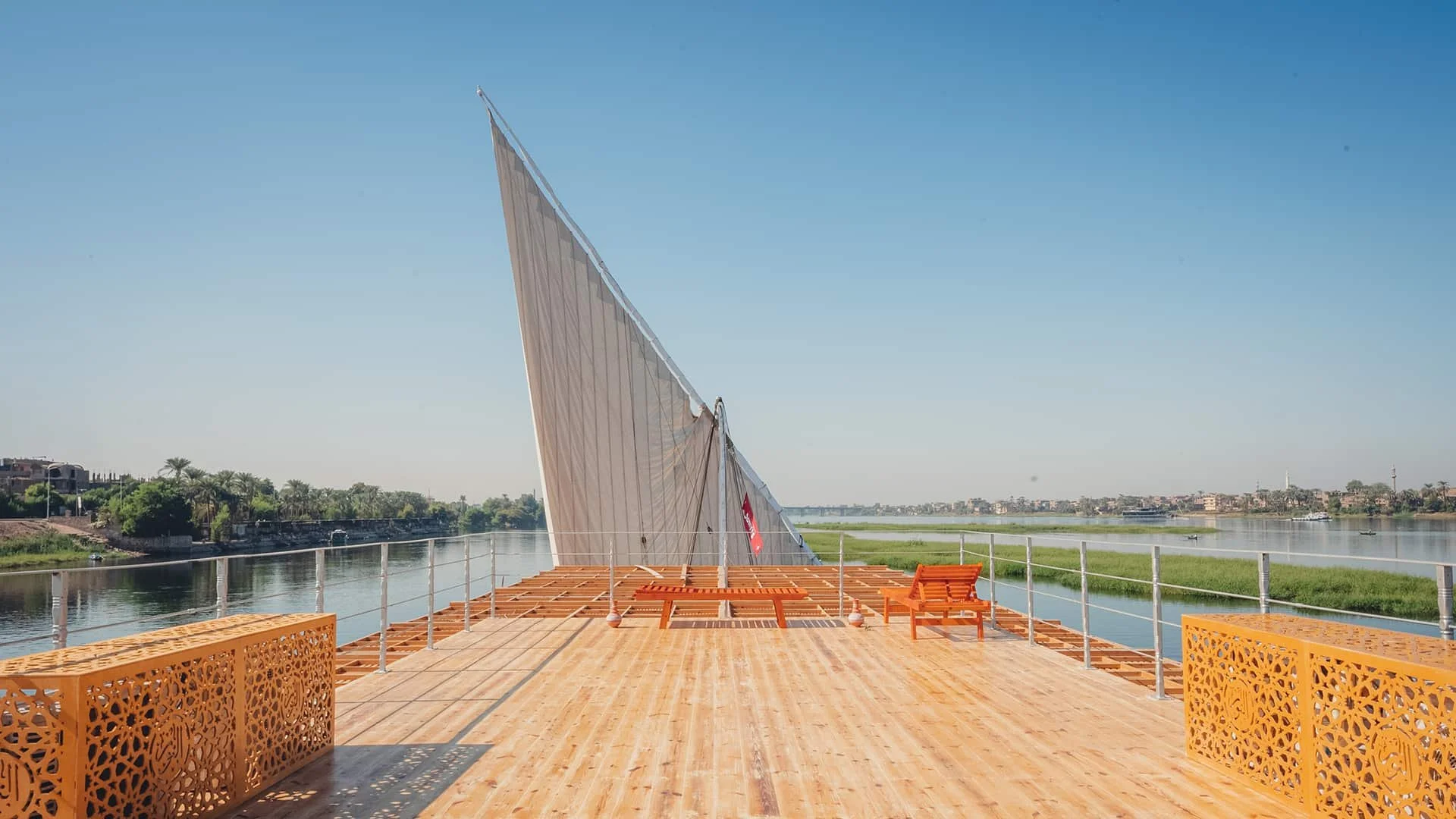 A boat with a large sail docked on a wooden platform on a river, with water and greenery in the background under a clear blue sky.