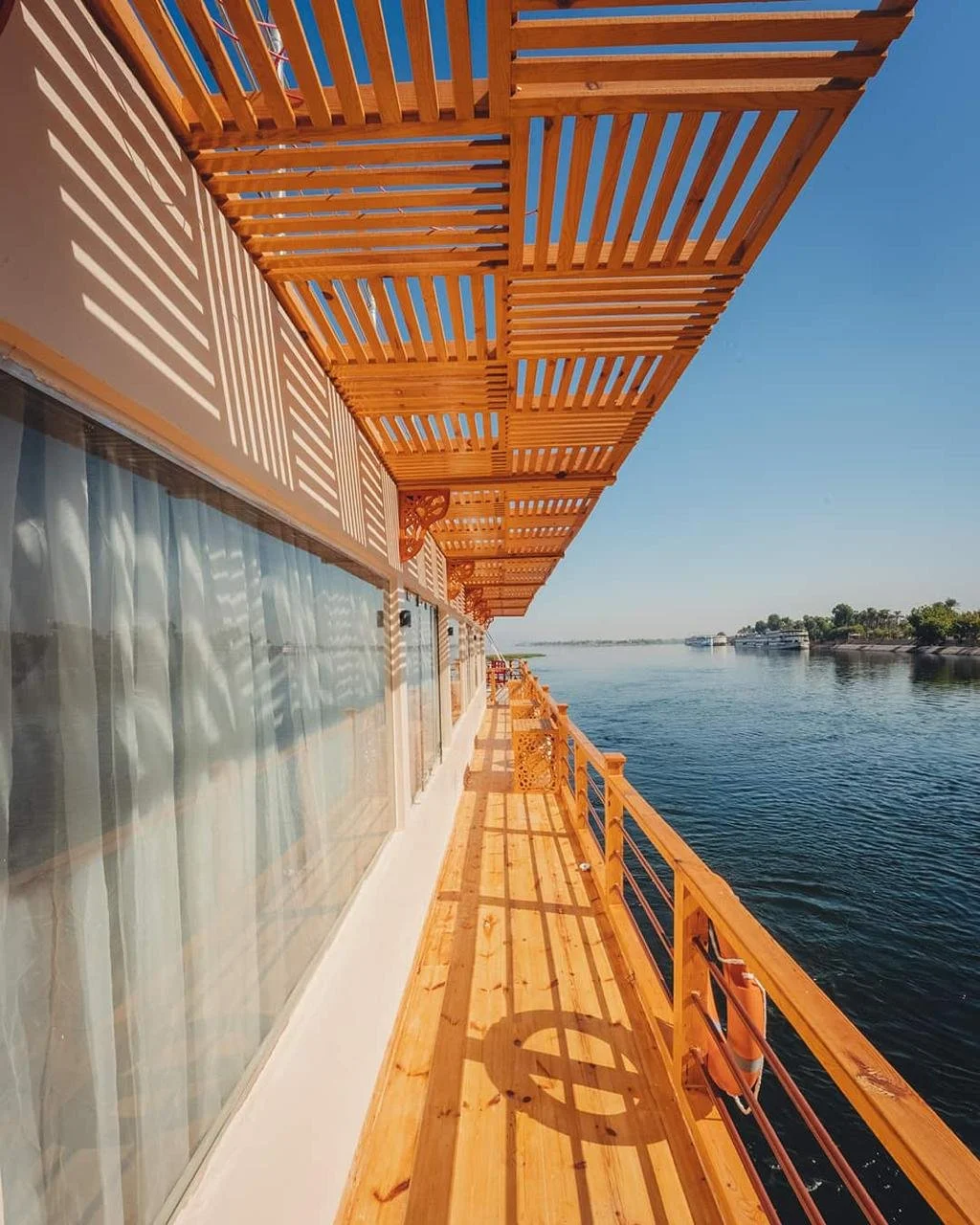 A view from a wooden balcony with slatted privacy screens, overlooking a river with boats and trees in the distance on a sunny day.