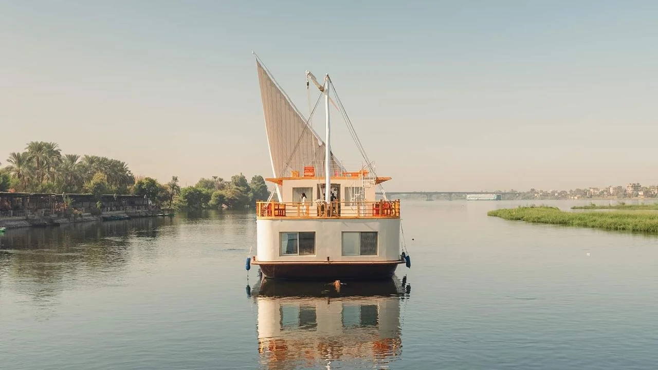 A houseboat on a calm river with trees and a distant bridge in the background during daytime.