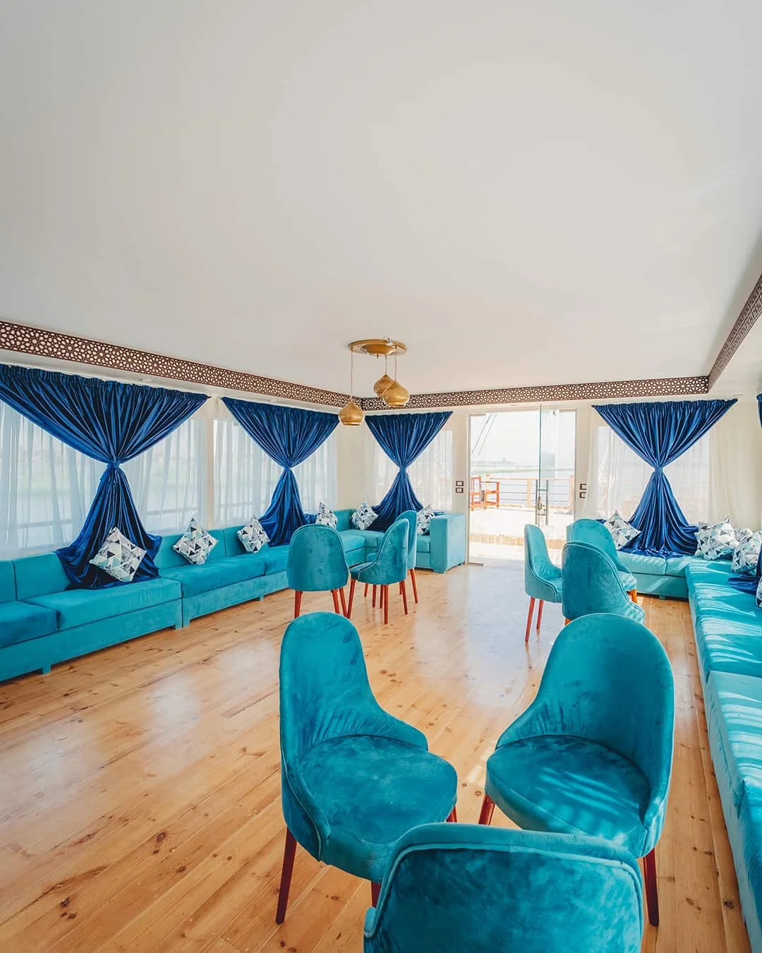 A spacious room with blue velvet chairs, blue curtains, and a wooden floor, with large windows and a balcony door.