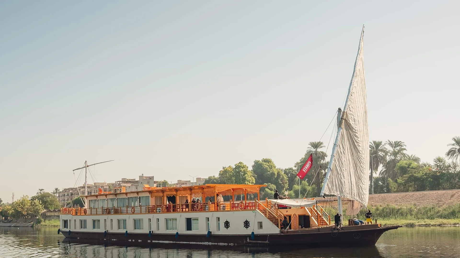 A large traditional boat with a white body and wooden accents on a calm river, with people aboard, and a tall white sail on the right side of the boat. Palm trees and buildings are visible in the background.