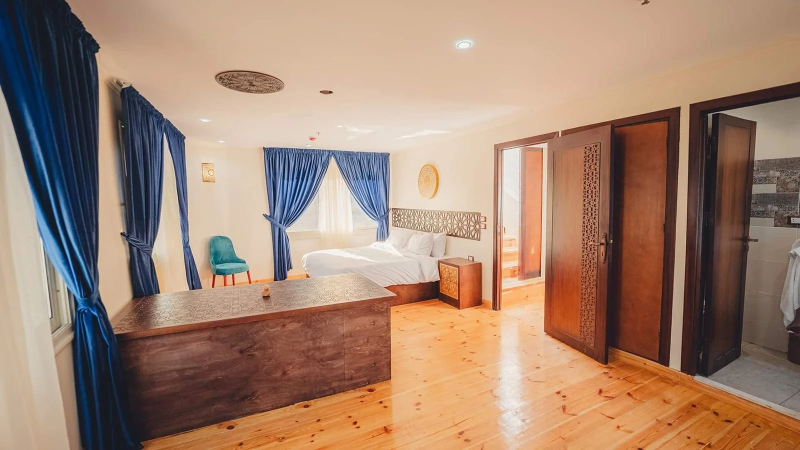 A hotel room with a bed, blue curtains, a turquoise chair, wooden floors, and a bathroom door open to reveal a tiled bathroom.