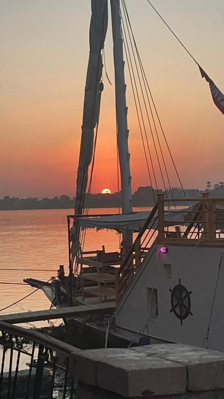 A sailboat docked at a pier during sunset with the sun partially visible on the horizon, creating an orange and pink sky reflection on the water.