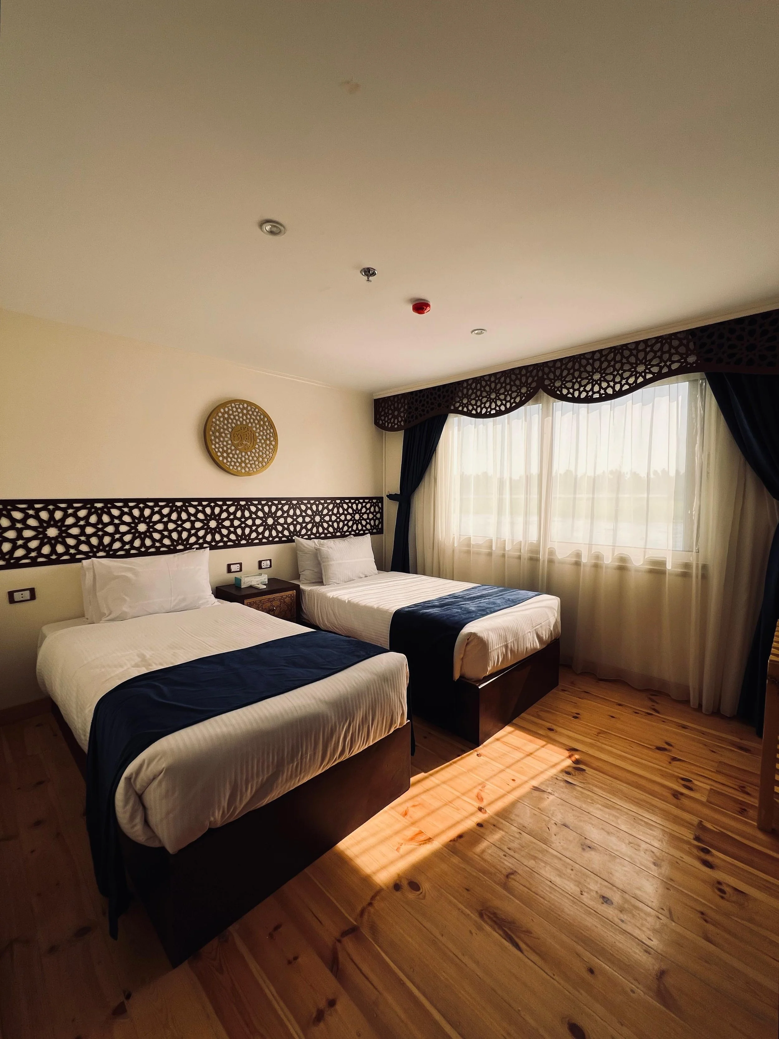 A hotel room with two single beds, dark wood headboards with geometric patterns, a bedside table, a window with sheer curtains and dark drapes, and a wooden floor.