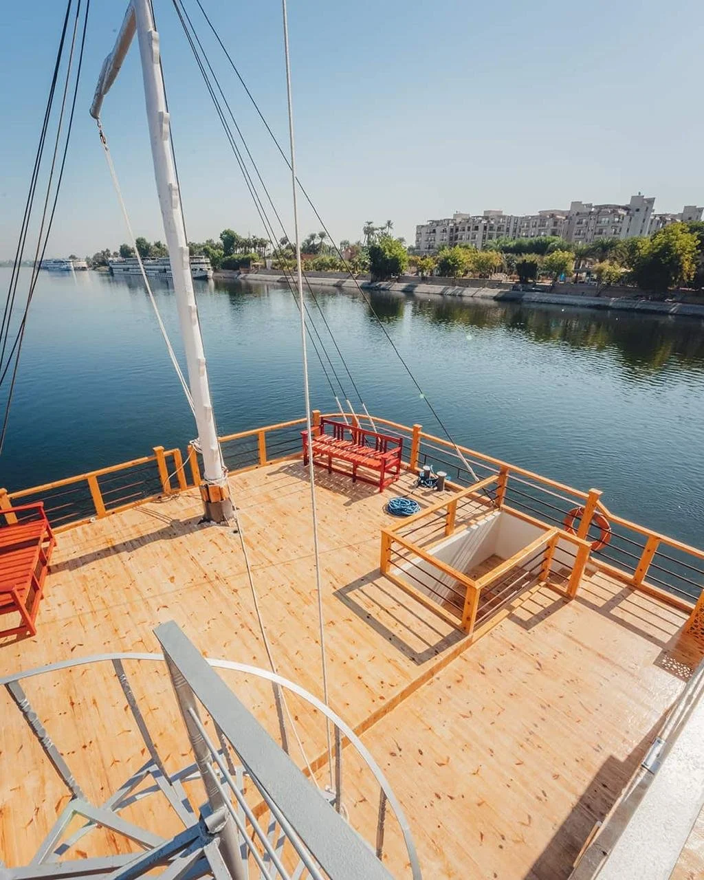 View of a boat deck with wooden flooring, red benches, railings, and a sailboat's mast, overlooking a river with buildings and trees in the background on a clear, sunny day.
