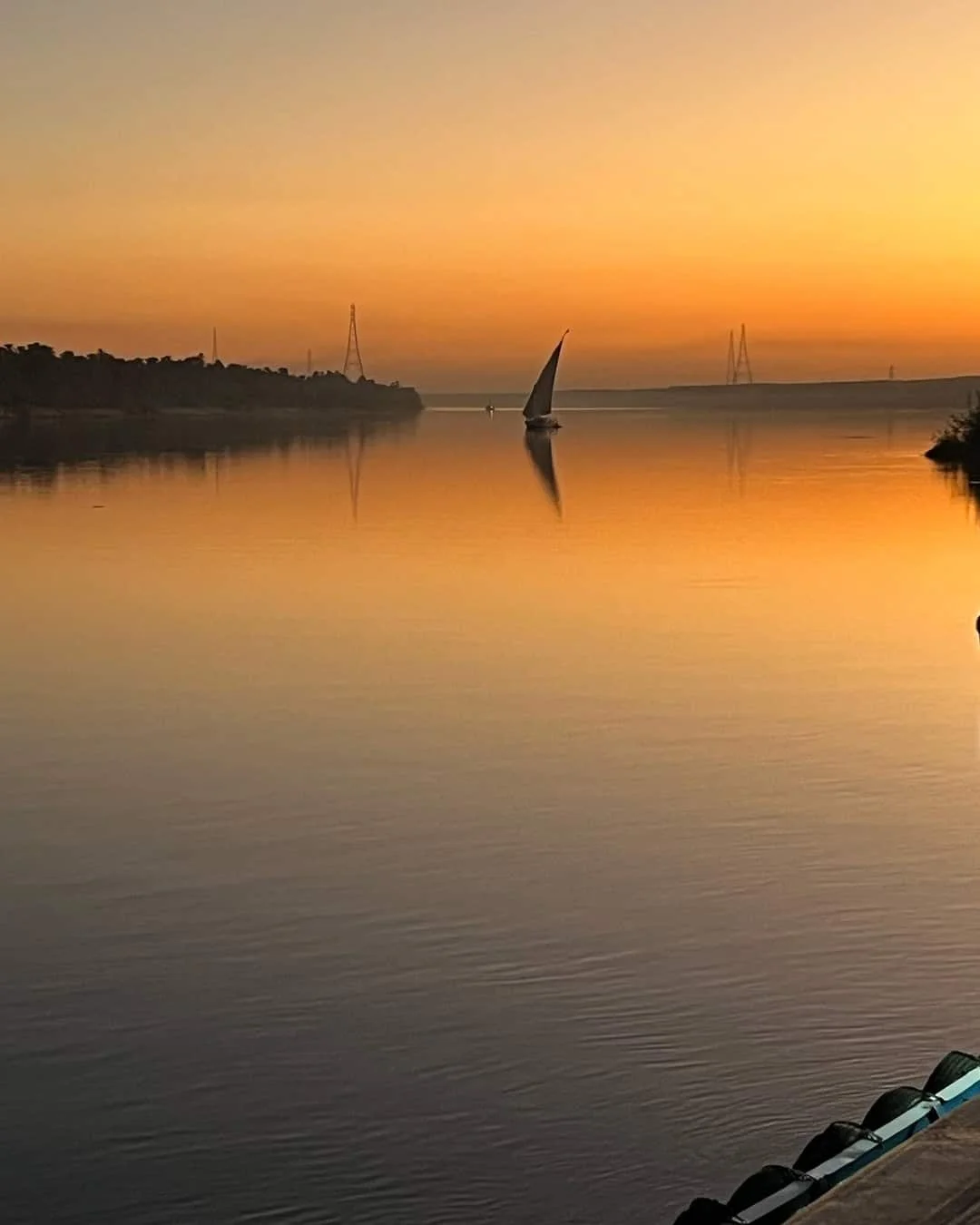 A calm river at sunset with a sailboat in the center, reflections on the water, and a bridge in the background.
