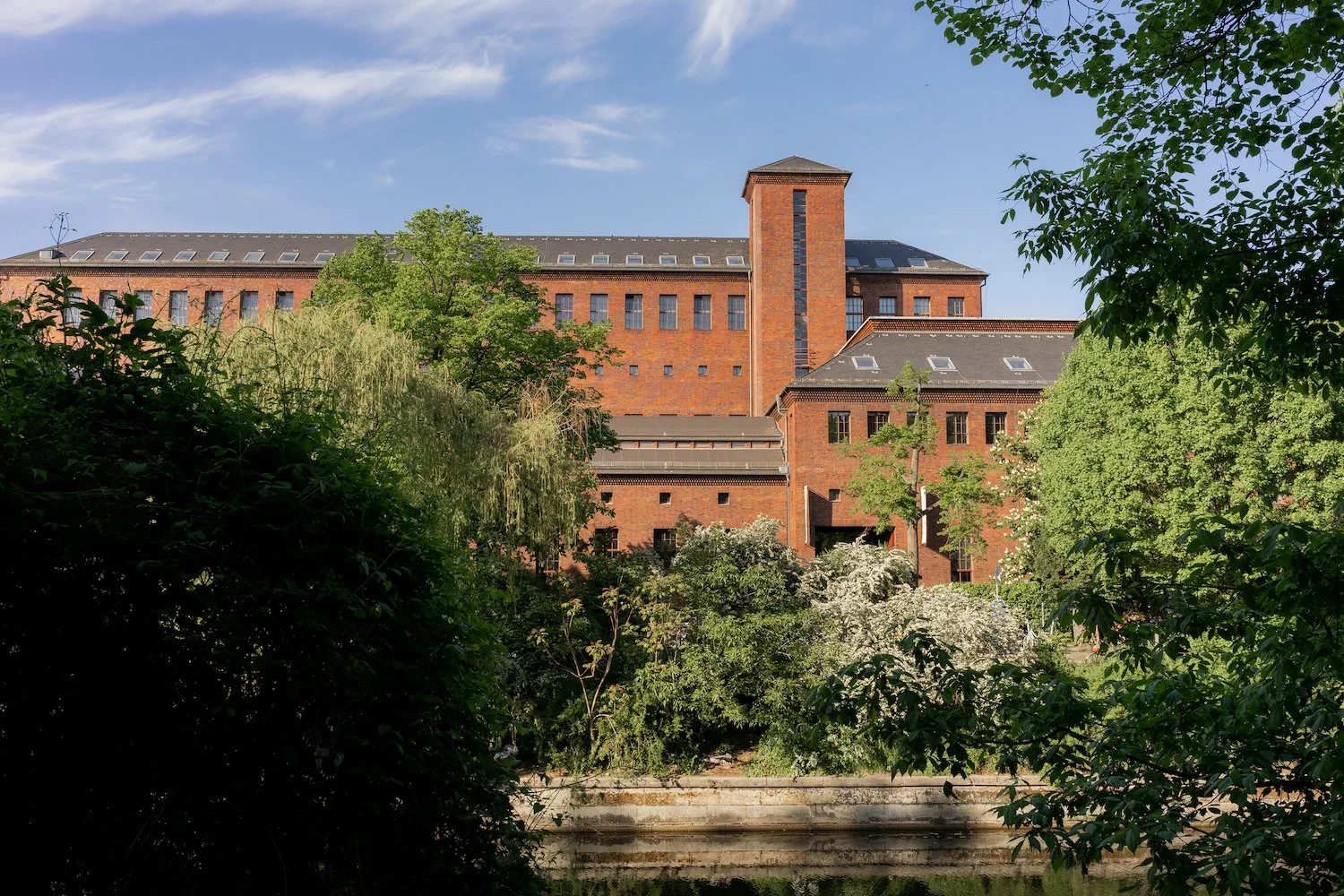 Das Umspannwerkgebäude in der Totale vom anderen Kanalufer aus fotografiert. Viele grüne Bäume und Büsche im Vordergrund.