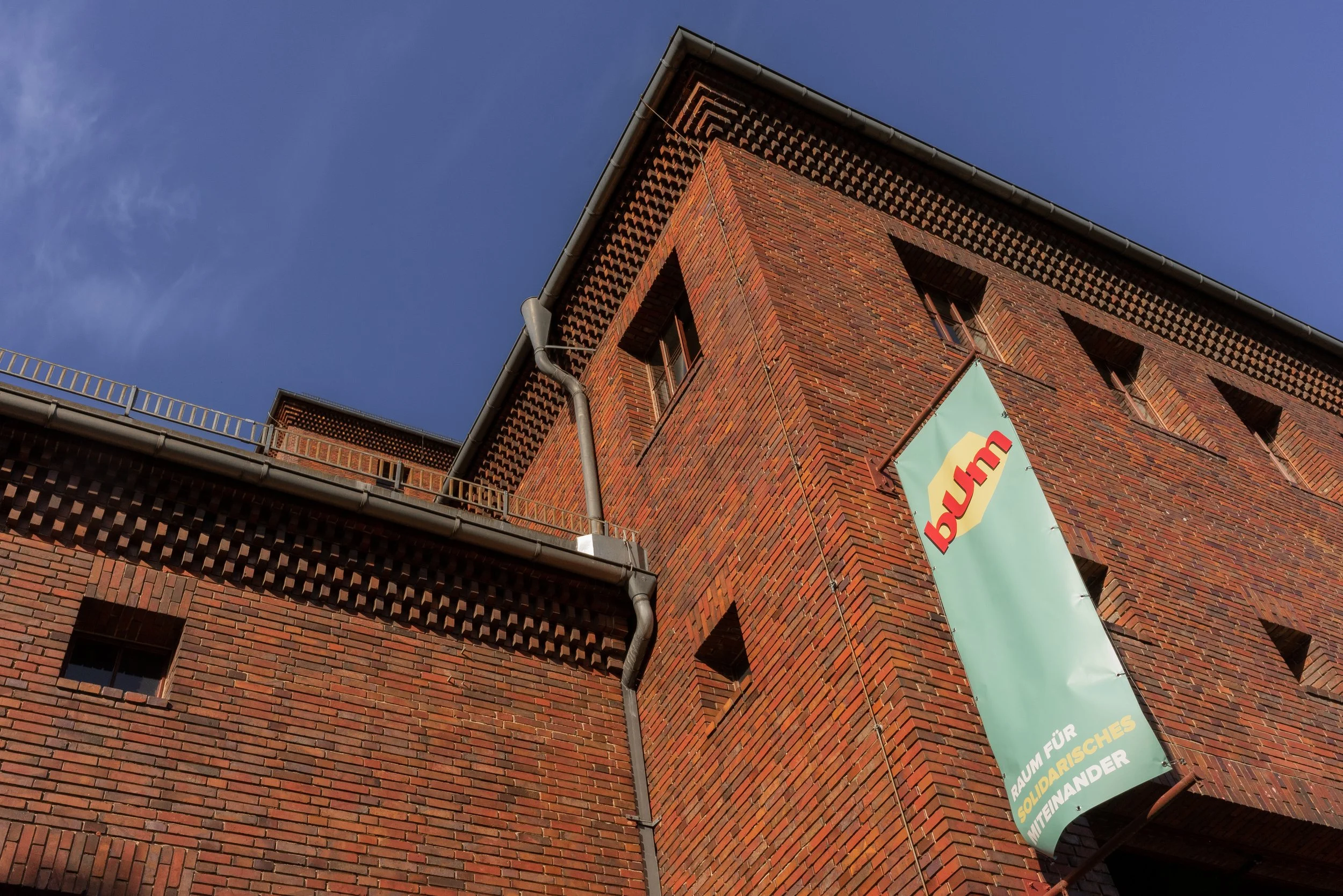 Ein Ausschnitt der bUm Fassade. Ein Backsteingebäude mit einem türkisblauen Banner mit dem bUm Logo.