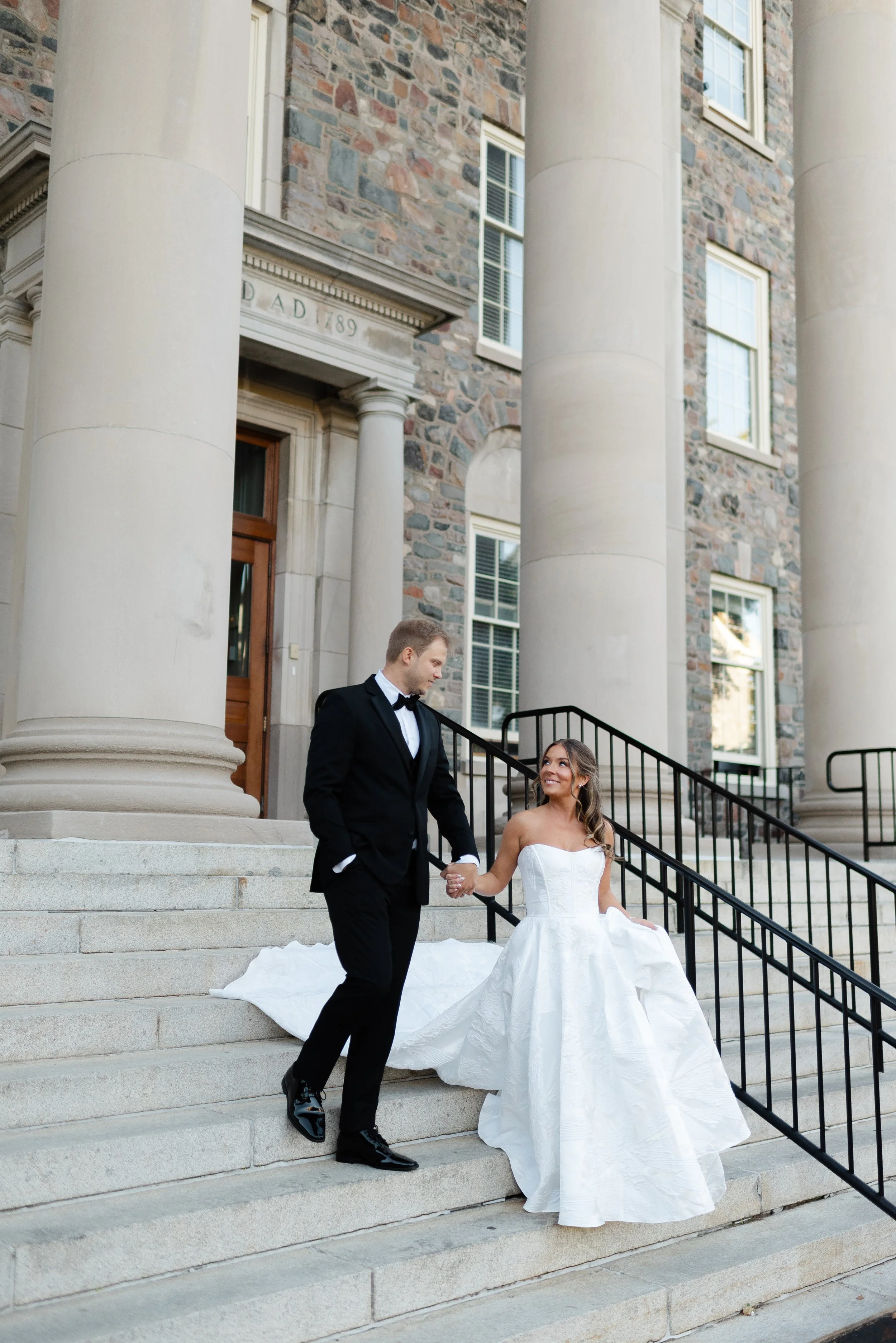 A bride and groom walking down the steps of a historic building with large columns, holding hands and smiling at each other in wedding attire.