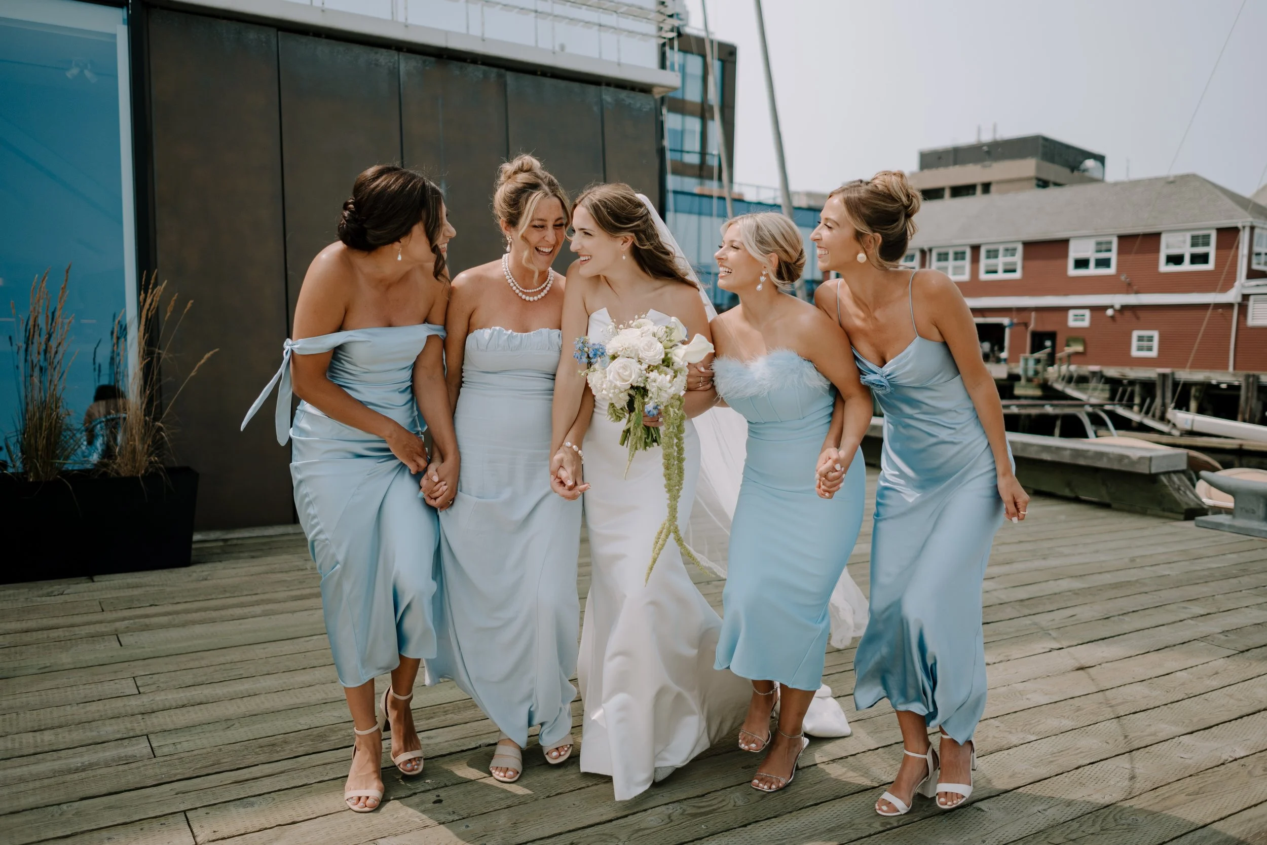 A bride in a white dress walking with her bridesmaids in light blue dresses on a wooden dock with boats and buildings in the background.