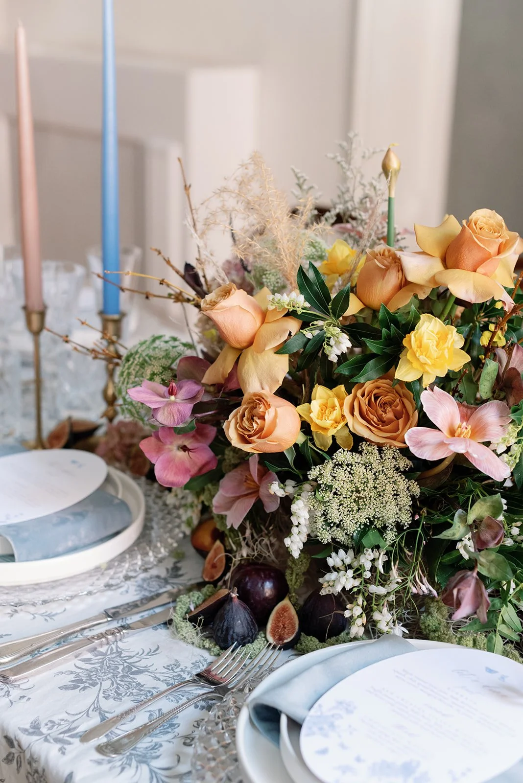 A floral centerpiece with peach, yellow, and pink flowers, figs, and greenery on a dining table set with silverware, plates, and a white napkin, with pink and blue candles in the background.