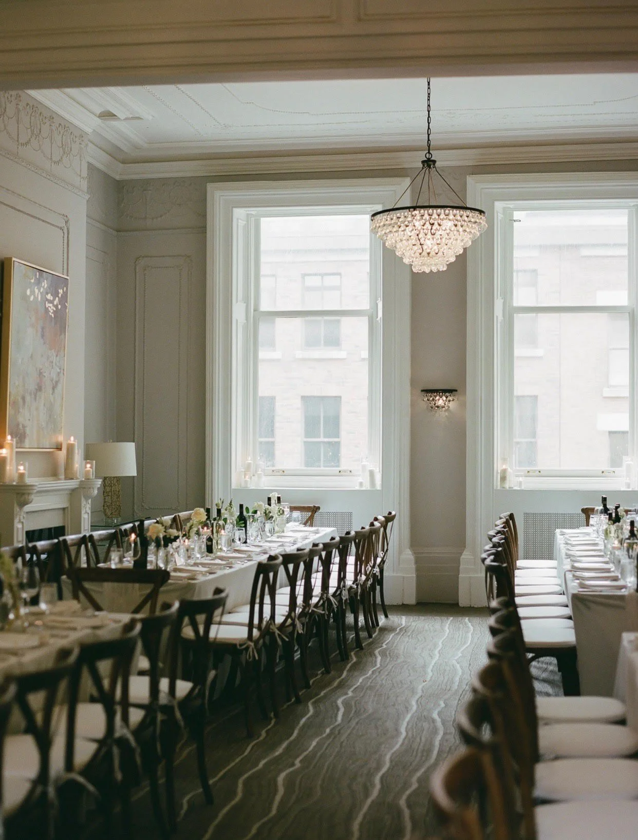 A dining room with two long tables set for a meal, adorned with candles, wine bottles, and glassware, in front of large windows with white frames, decorated with chandeliers and wall sconces, and a patterned carpet on the floor.
