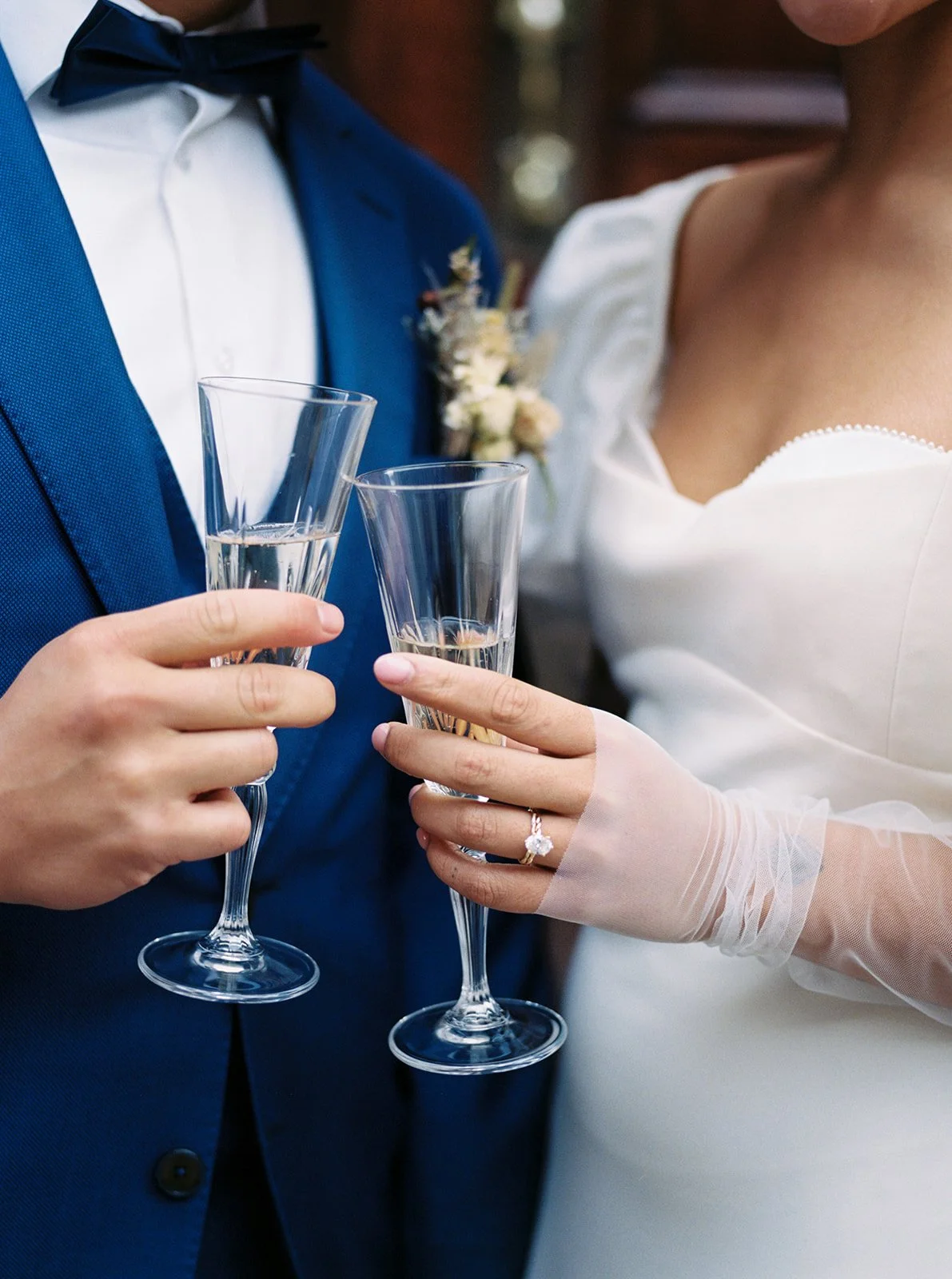 Close-up of a bride and groom holding champagne flutes during a celebration, possibly a wedding, with the bride wearing a ring and veil, and the groom in a blue suit.