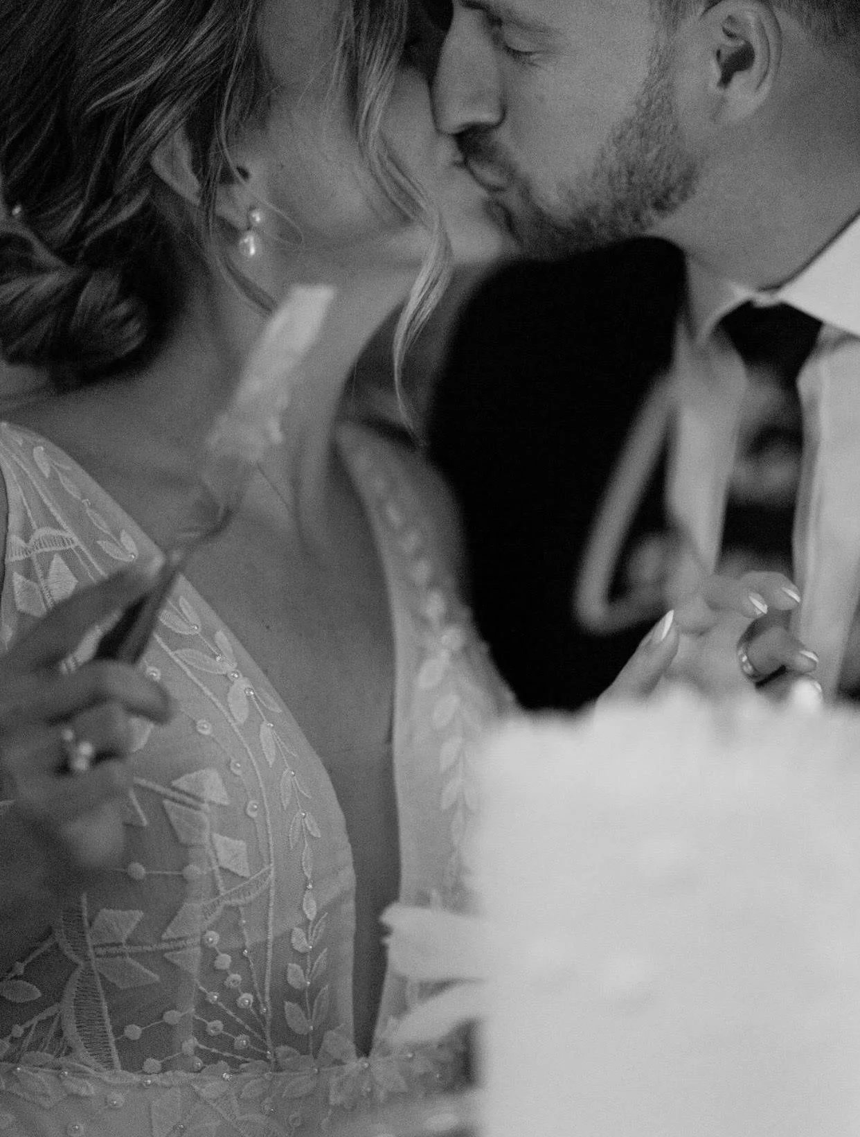 A black and white photo of a couple sharing a kiss at their wedding, with the bride in a lace dress and the groom in a suit and tie.