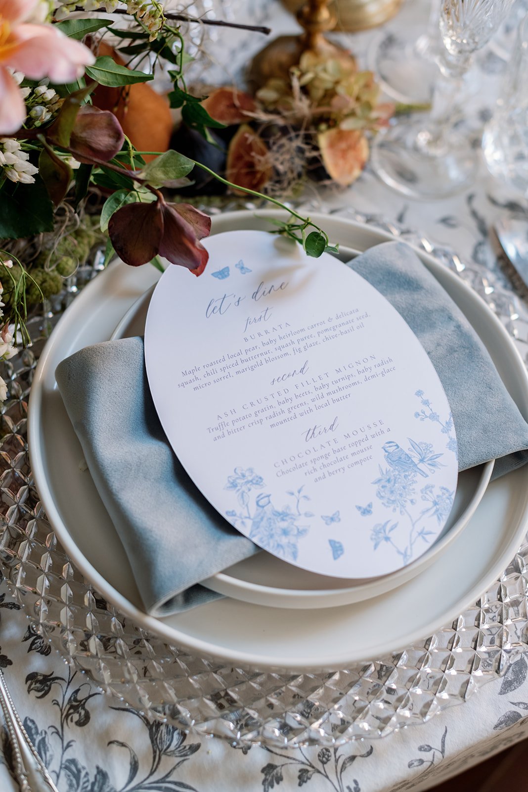 Elegant table setting with a white plate, a folded light blue napkin, a menu card, and surrounding floral decorations. The menu features three courses: First, BURRATA; second, ASH CRUSTED FILET MIGNON; third, CHOCOLATE MOUSSE, with decorative floral and butterfly illustrations.