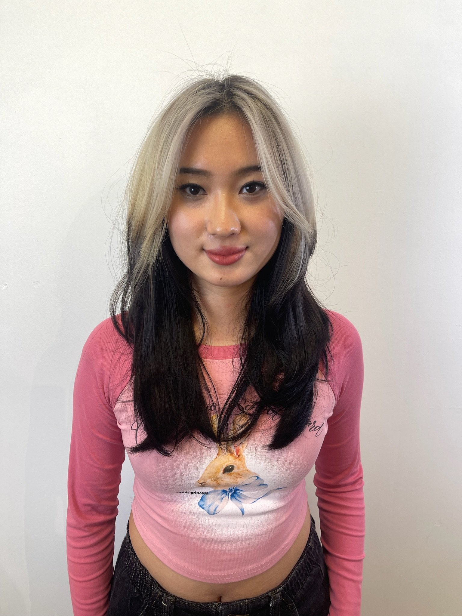 Woman with long, layered black and blonde hair, wearing a pink long-sleeve shirt with a rabbit graphic.