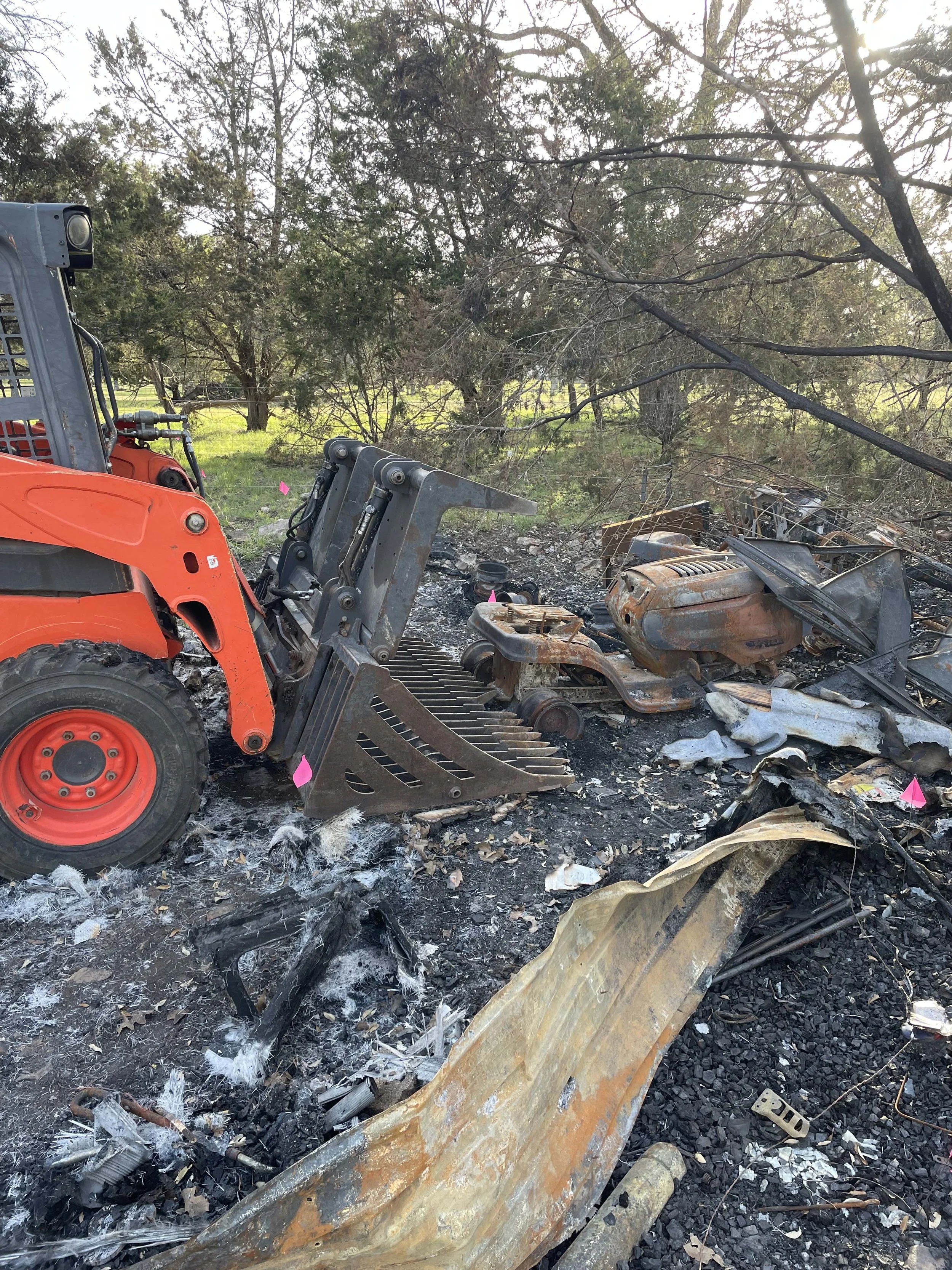 Burned and destroyed debris from a crash site, including parts of a vehicle, ash, and a skid-steer loader.