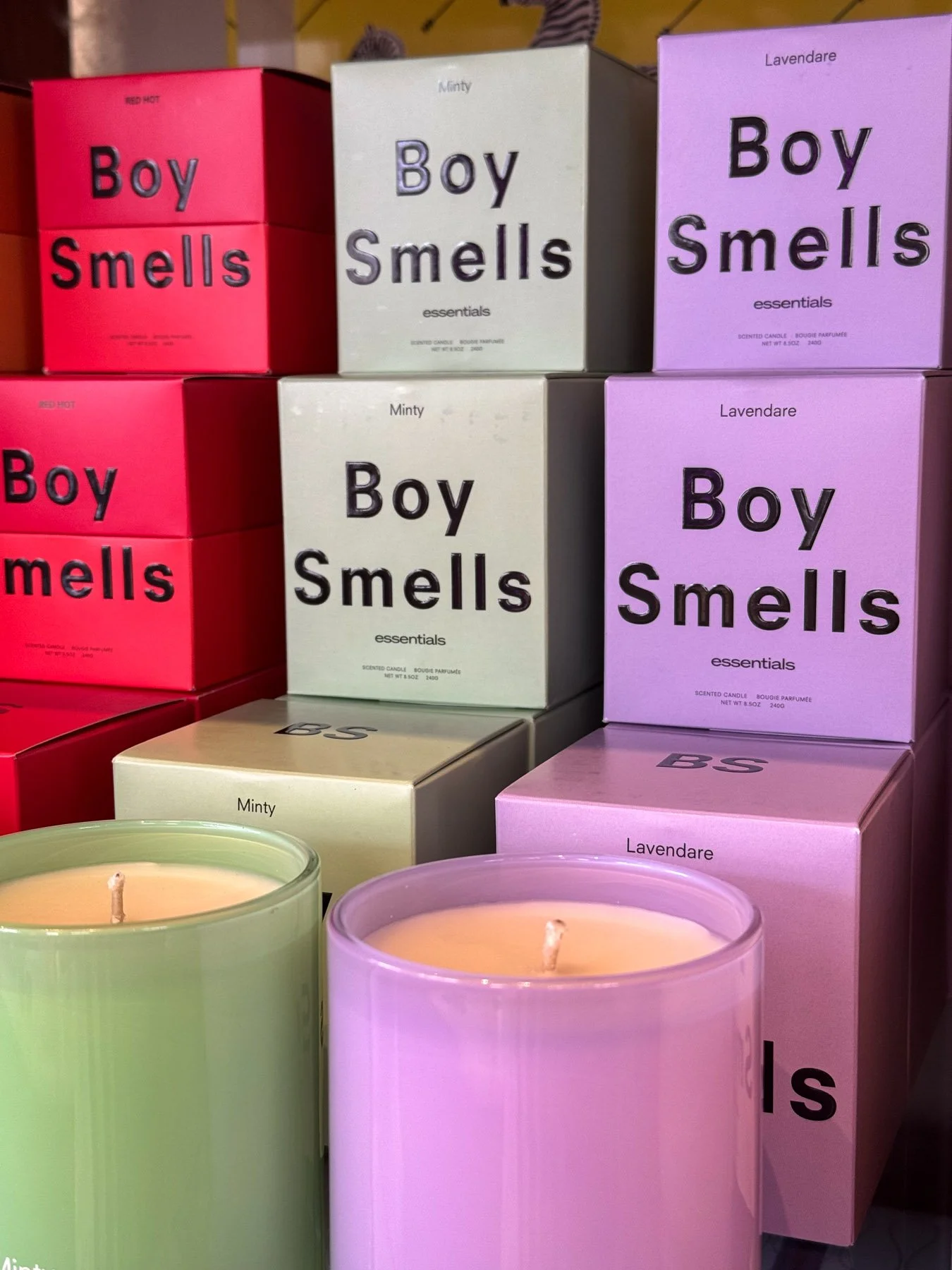 Variety of scented candles with boxes labeled "Boy Smells" in different colors including red, white, and purple, with candles in front of the boxes in matching colors.