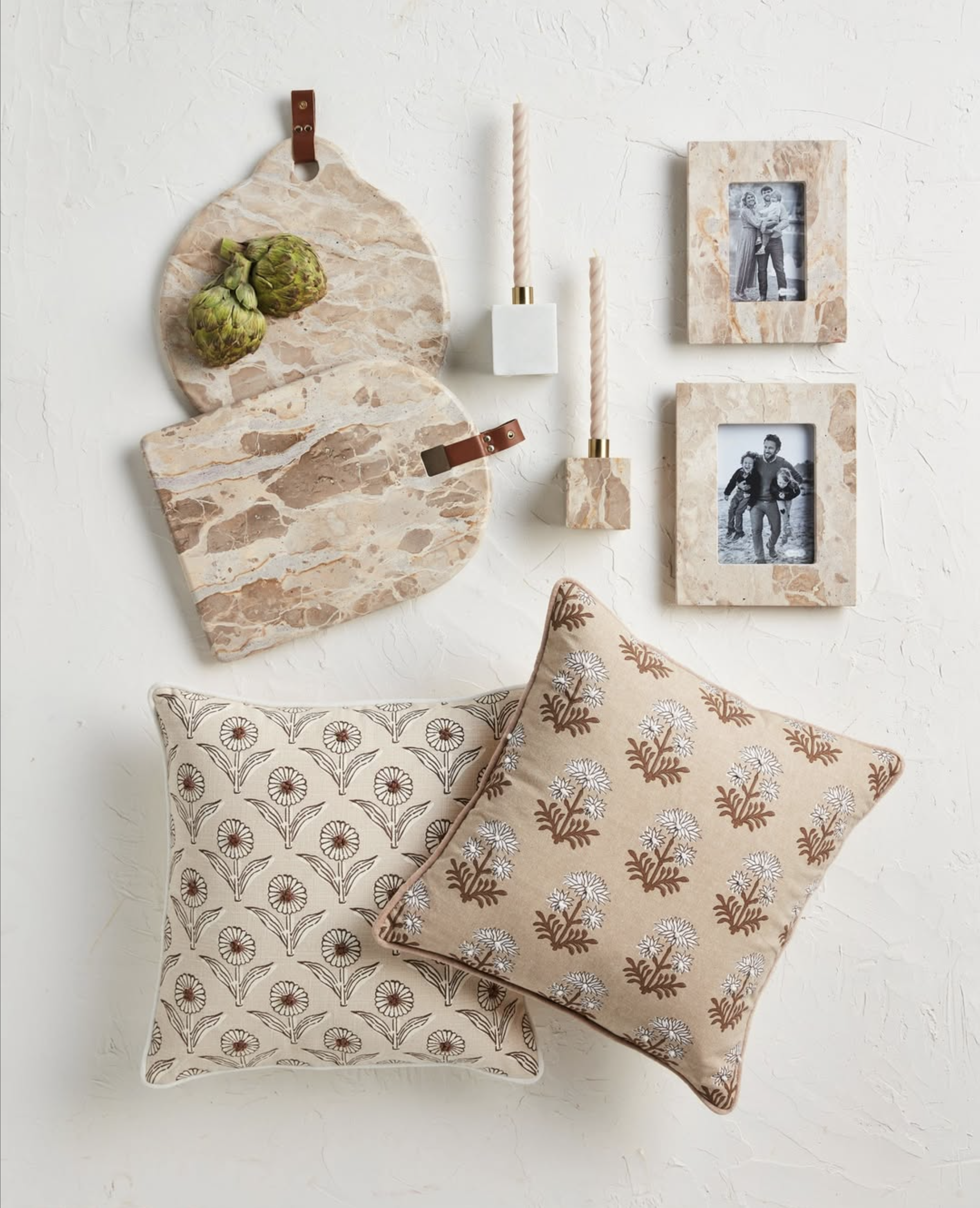 Two decorative pillows with floral patterns, two marble photo frames with black and white photos, two marble candle holders with spiral candles, and two marble cutting boards with artichokes on a textured white surface.