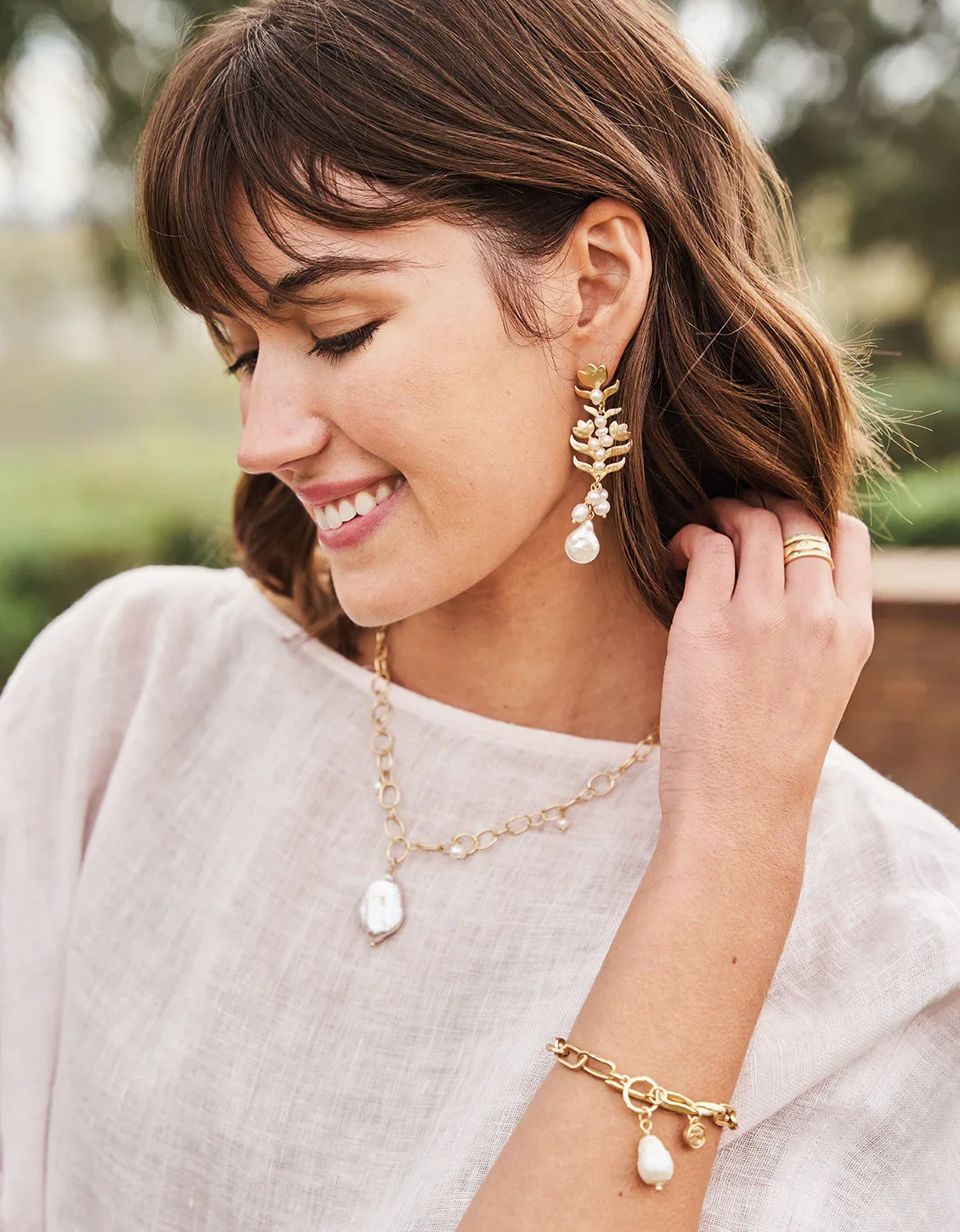 A woman with short brown hair smiling and wearing gold jewelry, including earrings, a necklace, a bracelet, and rings, outdoors with a blurred background.