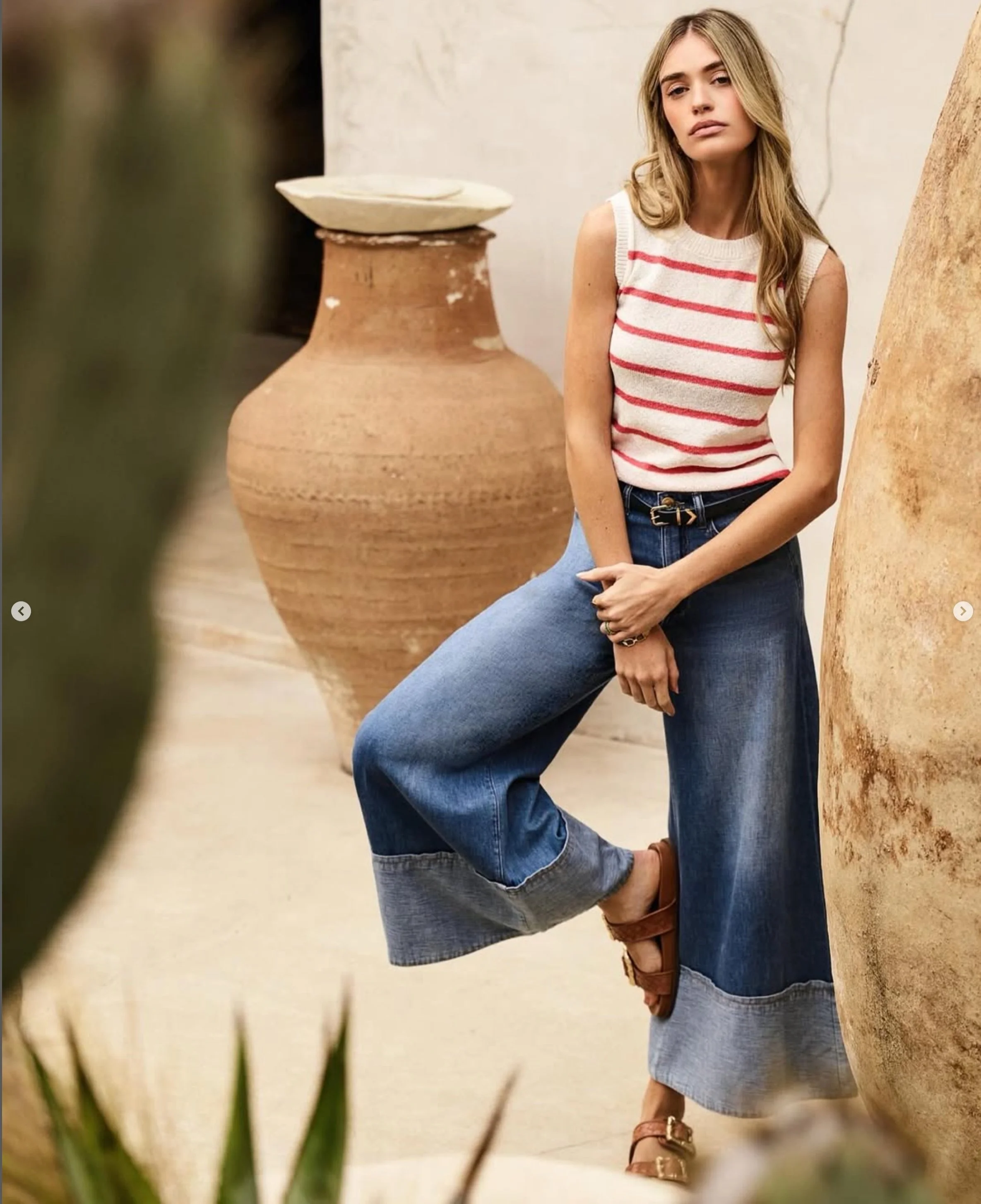 Woman in sleeveless striped top and wide-leg jeans leaning against a large ceramic pot, with another pot in the background, indoors.