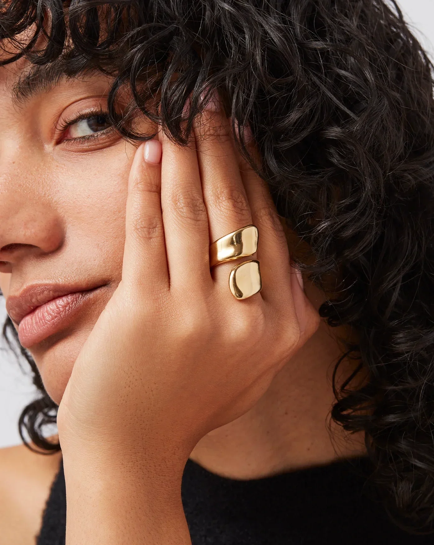 Close-up of a woman's face and hand with gold jewelry, curly hair, and a black top.