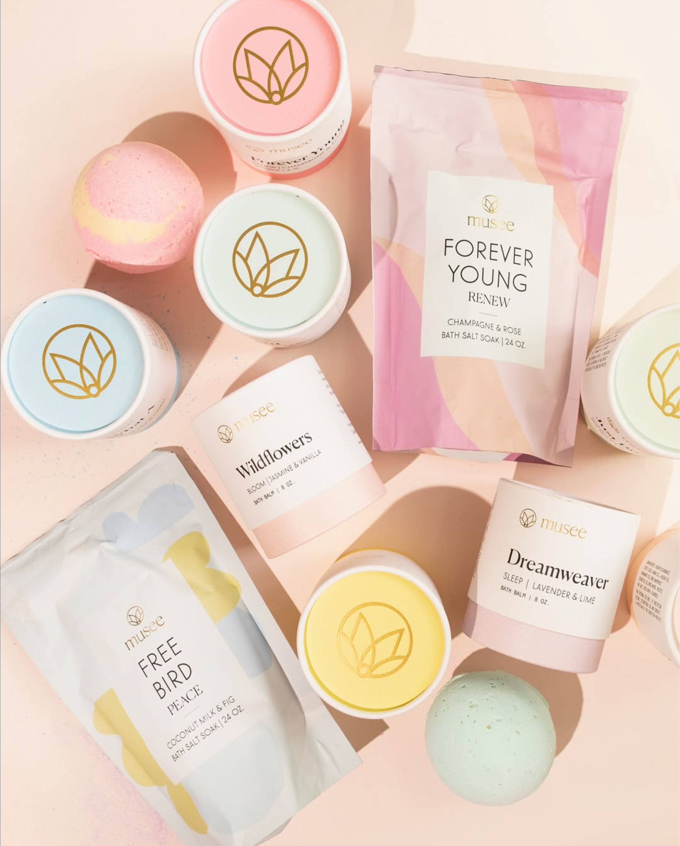 Assorted bath products with pastel packaging, including bath salts, bath balm, and bath bombs from the Musee brand, arranged on a pink surface.